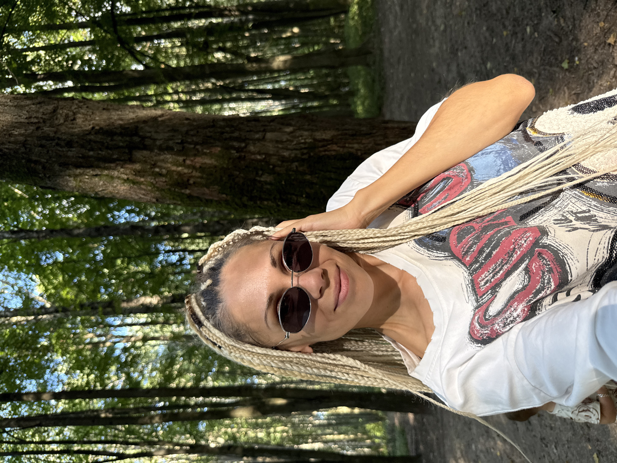A woman with blonde braids and sunglasses is taking a selfie in a forest. She is wearing a white t-shirt with a graphic design. The background shows tall trees and dappled sunlight.