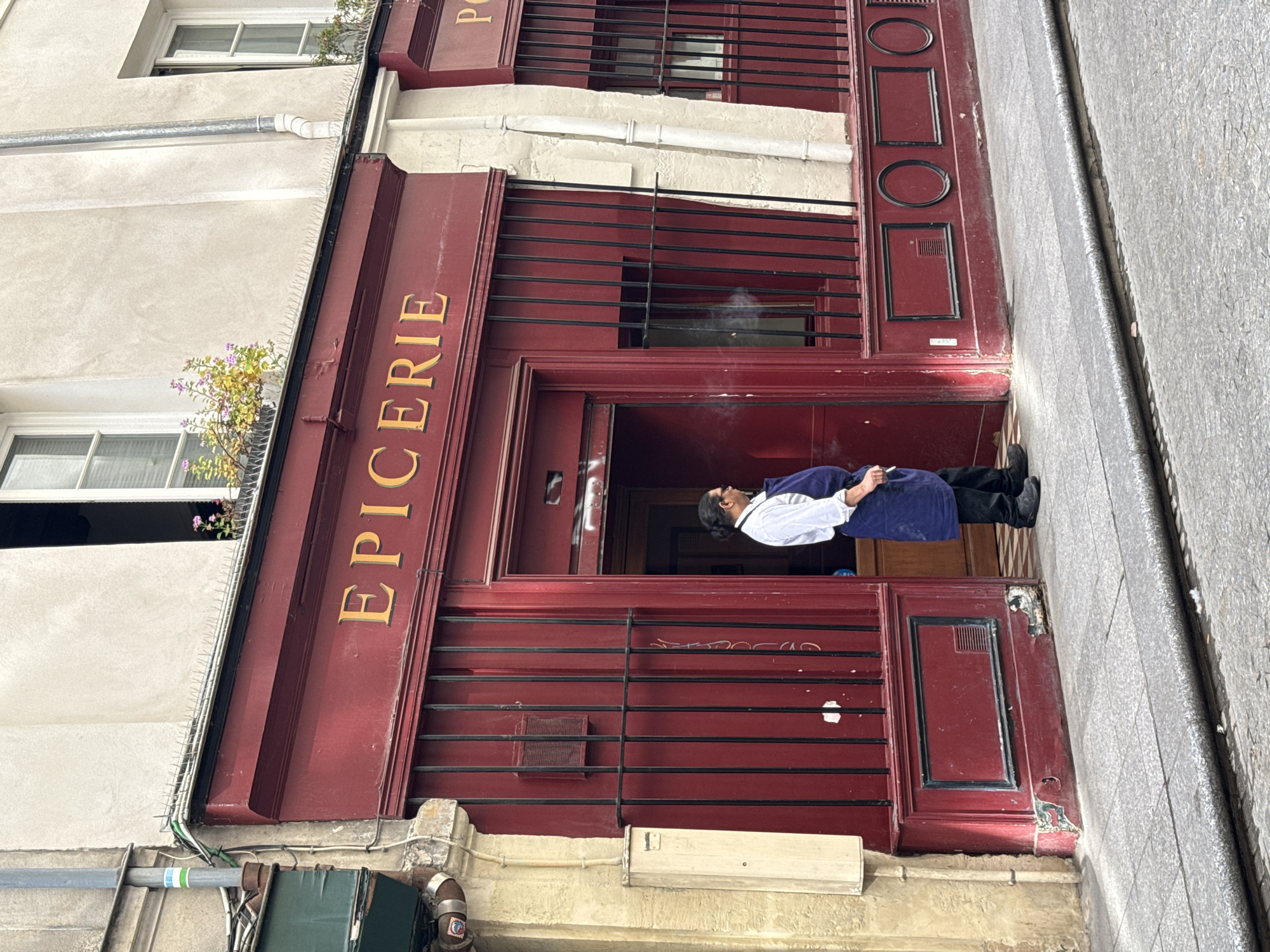 A man stands in the doorway of a small shop in Paris, France. The shop's façade is painted a deep red color. Above the doorway, the word "EPICERIE" is written in gold letters. The man is wearing a white shirt, a blue apron, and black pants. He is holding a cigarette in his hand and appears to be smoking. The street outside the shop is paved with cobblestones.