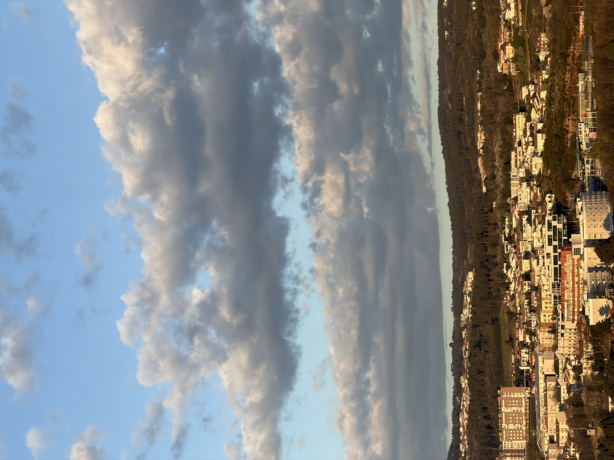 The image shows a vertical view of Vila Real, Portugal, at sunset. The sky contains patches of blue with intermittent gray and white clouds. Below the horizon, the city is lined with buildings and trees.
