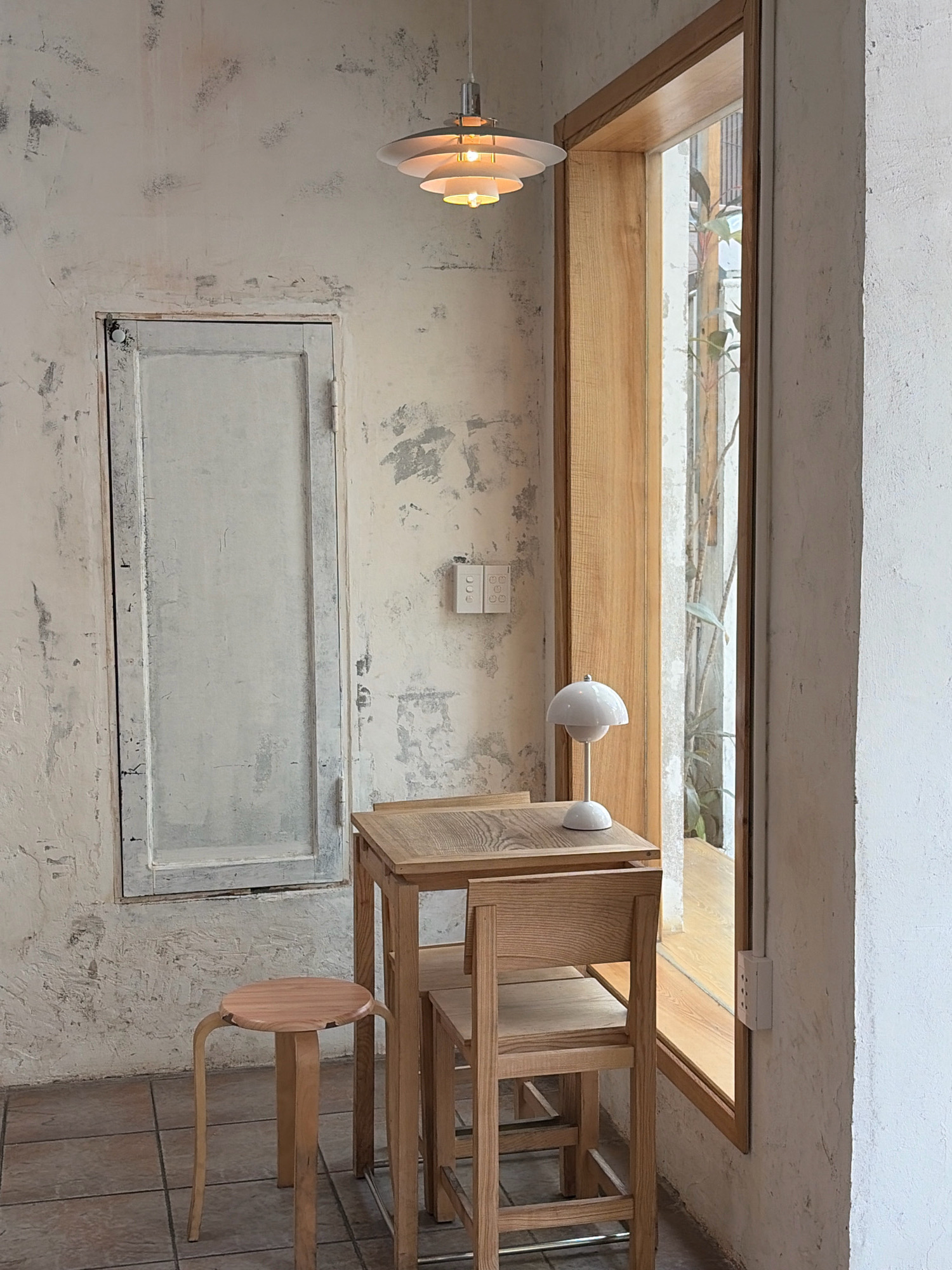 A minimalistic cafe interior in Da Nang, Vietnam, features a distressed white wall with subtle gray markings, a vertical panel resembling a closed door, and a large wooden-framed window. To the right of the window, a small wooden table is set with a white desk lamp and a wooden chair. A round wooden stool stands to the left of the table. A modern pendant light hangs from the ceiling. The floor is tiled in a muted brown. Plants are visible through the window.