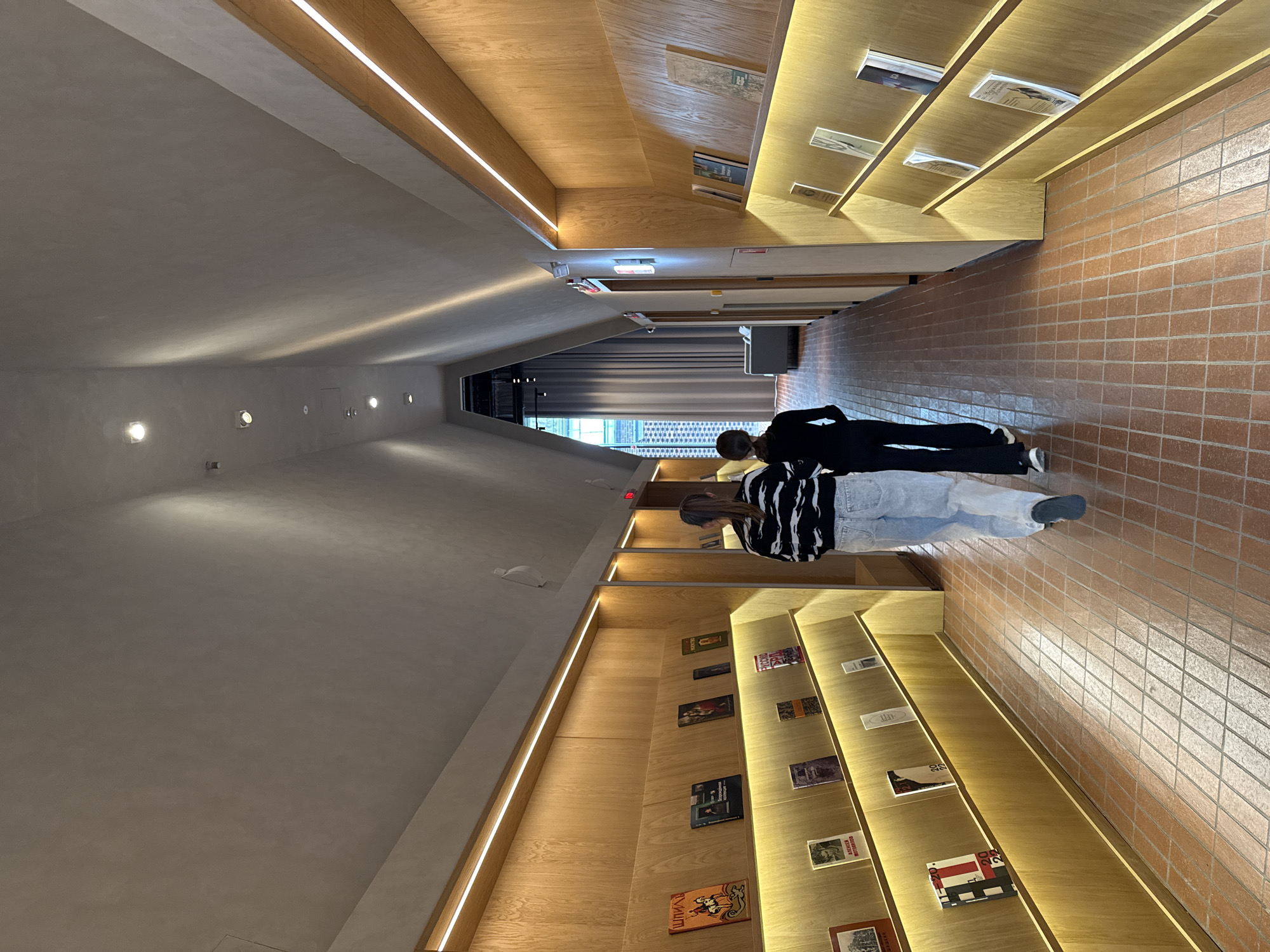 A slightly high-angle, interior shot depicts two individuals in what appears to be a bookstore or library setting. The overall architecture is modern, featuring a converging ceiling and walls that give a sense of perspective leading toward the back of the building.
On the right, a wall is constructed of brick-like tiles. On the left, the wall appears to be a smooth, grey surface. Both walls feature wooden shelves containing various books and other items. The shelves are illuminated by subtle, integrated lighting.
Two people are present. One person, dressed in all black, is leaning over. Another person, wearing a black and white striped shirt and blue jeans, is standing closer to the camera.
The setting could be the Perm Central City Library in Perm, Russia. The architectural style, with its sloped ceilings and integrated lighting, is consistent with the design of modern libraries and bookstores.