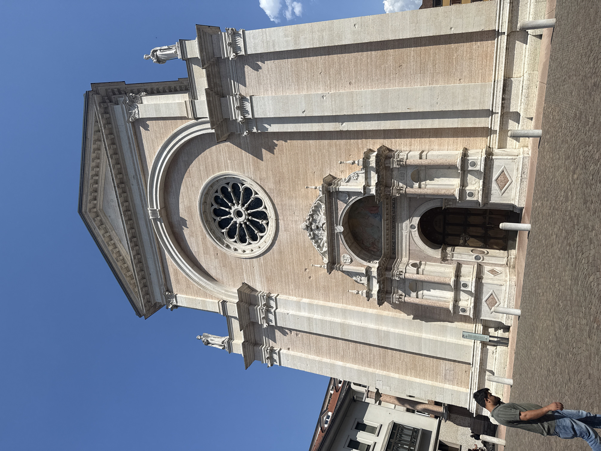 This is a full, eye-level, outdoor, slightly low-angle, wide shot photograph of the exterior of the Duomo of Trento, also known as the Cathedral of Saint Vigilius, located in Trento, Italy. The imposing facade of the cathedral dominates the frame, constructed with bands of light pink and cream-colored stone. A large, ornate rose window is centrally positioned above the entrance. Flanking the rose window, at the upper corners of the facade, are two stone statues of figures. The entrance features a grand arched doorway made of dark wood, recessed within a highly decorated stone portal. Above the doorway, there is a lunette containing a fresco. The sky is a clear, vibrant blue, with a few scattered white clouds visible on the right side of the frame. In the foreground, a man in a dark olive-green t-shirt and blue jeans walks from left to right, looking down. The ground is paved with cobblestones. White bollards are spaced along the edge of the plaza. To the left of the cathedral, a portion of a multi-story building with windows and a dark balcony is visible. The sunlight casts distinct shadows on the cathedral's facade.