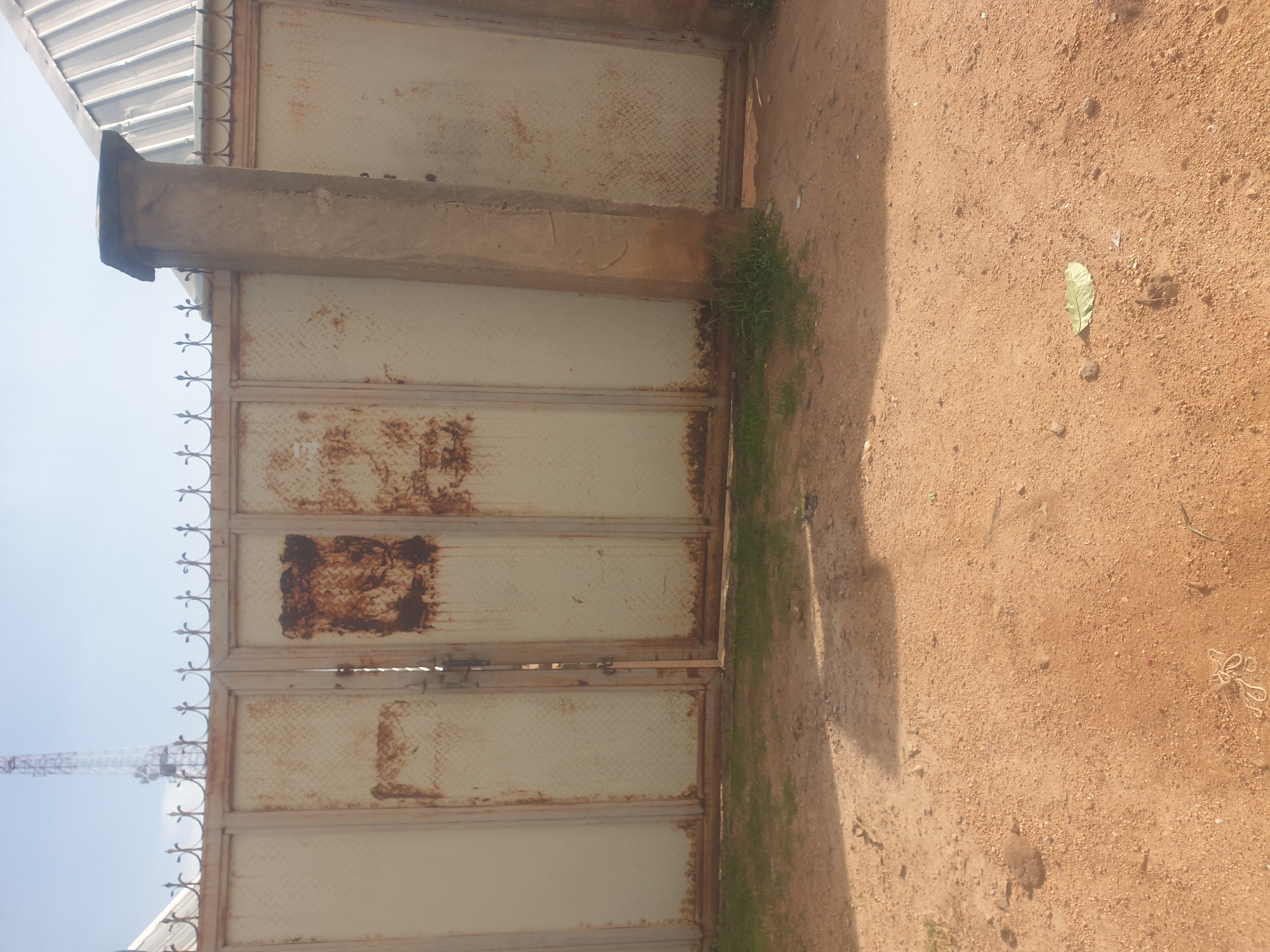 In Jauro Yinu, Nigeria, a weathered metal gate stands partially open, revealing a glimpse of what lies beyond. The gate, painted a pale color that has faded and rusted in places, is adorned with decorative spikes along its top edge. To the right, a sturdy concrete pillar supports a portion of the gate and the fence. The ground in front of the gate is a mixture of sandy soil and sparse grass, with a few scattered leaves and debris. In the background, a tall communication tower pierces the clear sky, suggesting the presence of infrastructure within the city. The overall scene is one of everyday life, with the gate serving as a boundary between public and private spaces.