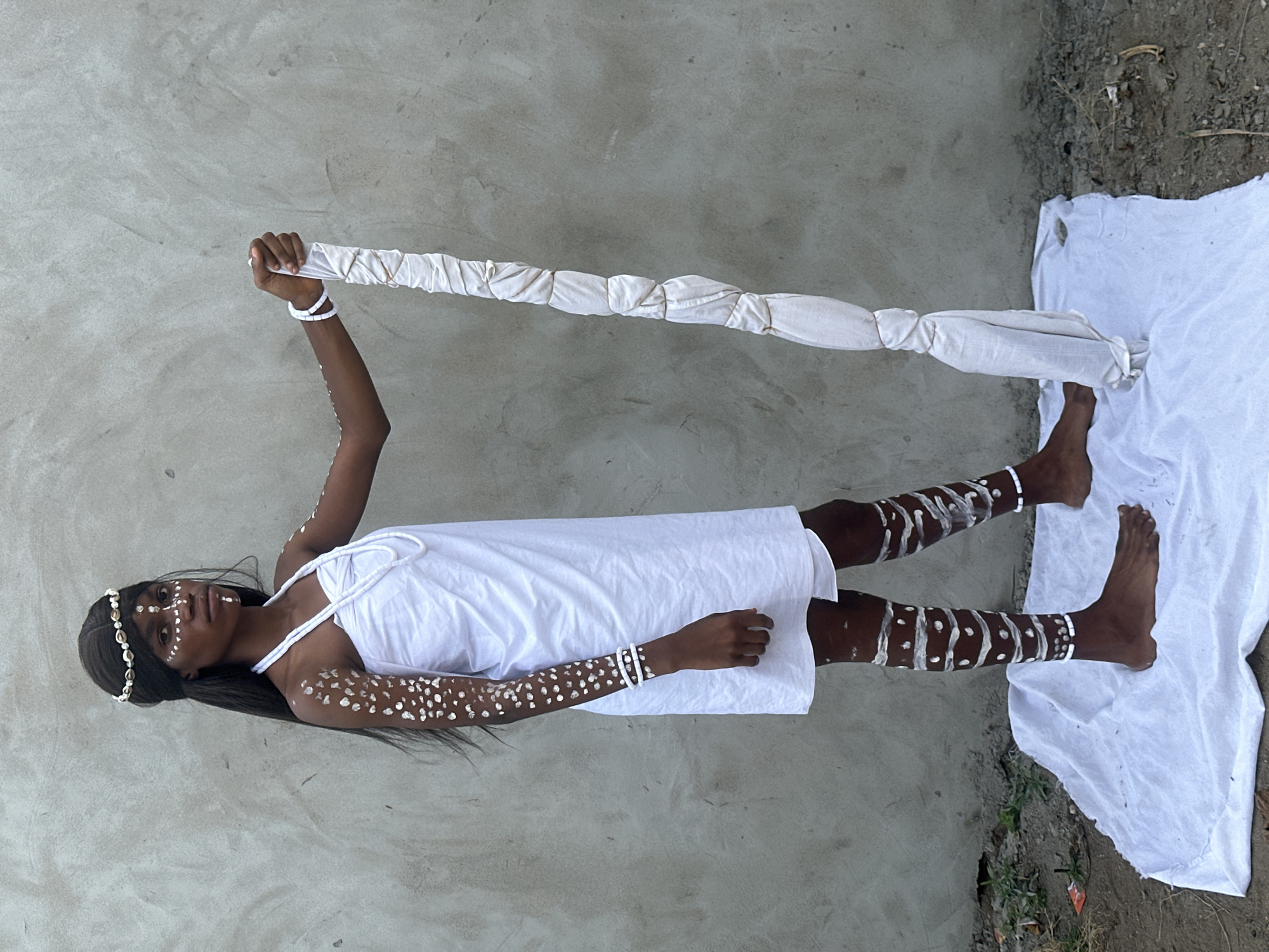 A young woman stands in front of a concrete wall in Badagry, Nigeria. She is adorned with white body paint, geometric patterns on her face and arms, and she wears a white dress and a seashell headband. She holds a long, white cloth staff in her right hand. Her feet are bare and resting on a white cloth spread on the ground. The ground is a mixture of dirt and gravel.