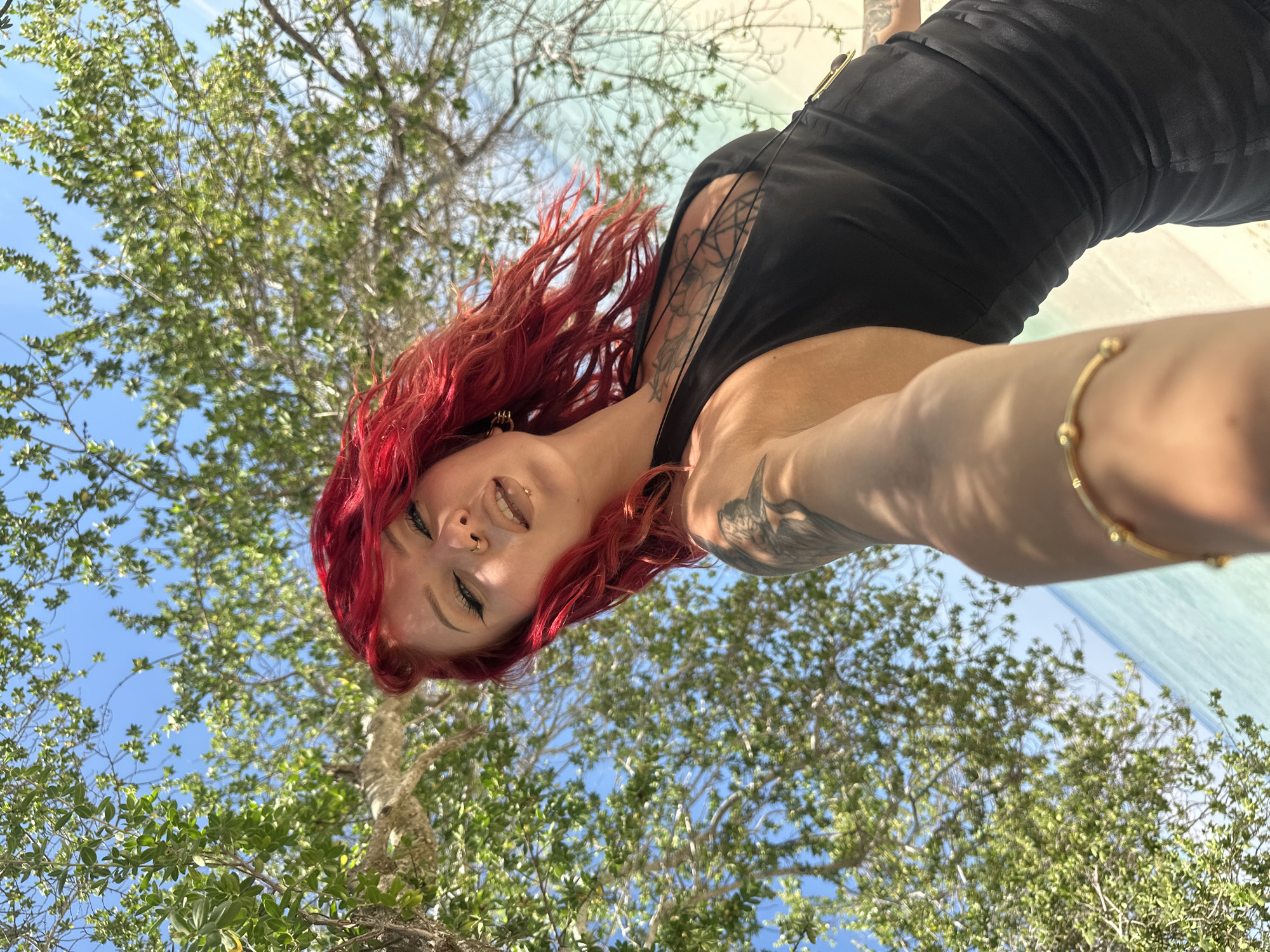 A woman with red hair is taking a selfie. She is wearing a black top. Her left arm is extended towards the camera, and a gold bracelet is visible on her wrist. Tattoos are visible on her neck and arm. Trees with green leaves and a blue sky are in the background. The location is not identifiable from the image.