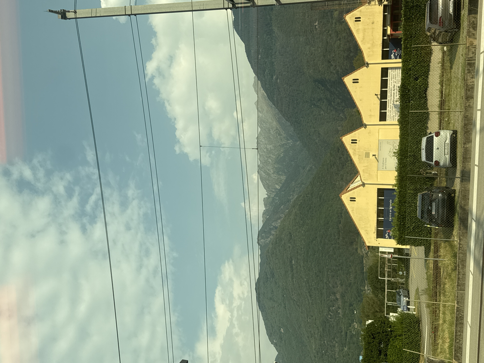 The image captures a daytime view from a moving vehicle, likely a train, looking towards a row of yellow industrial buildings with distinctive pointed roofs, situated in Bellinzona, Switzerland. The buildings appear to be warehouses or workshops, with signs indicating "Centro artigianale" (Artisanal Center). In front of the buildings, a neatly trimmed hedge separates them from a fenced area containing two parked cars: a white Volkswagen Golf and a darker colored SUV. Train tracks are visible in the foreground. Behind the buildings, a densely forested mountain range dominates the background, with a partly cloudy blue sky above. The overall scene depicts an industrial or commercial area on the outskirts of the city, backed by the natural beauty of the Swiss Alps.