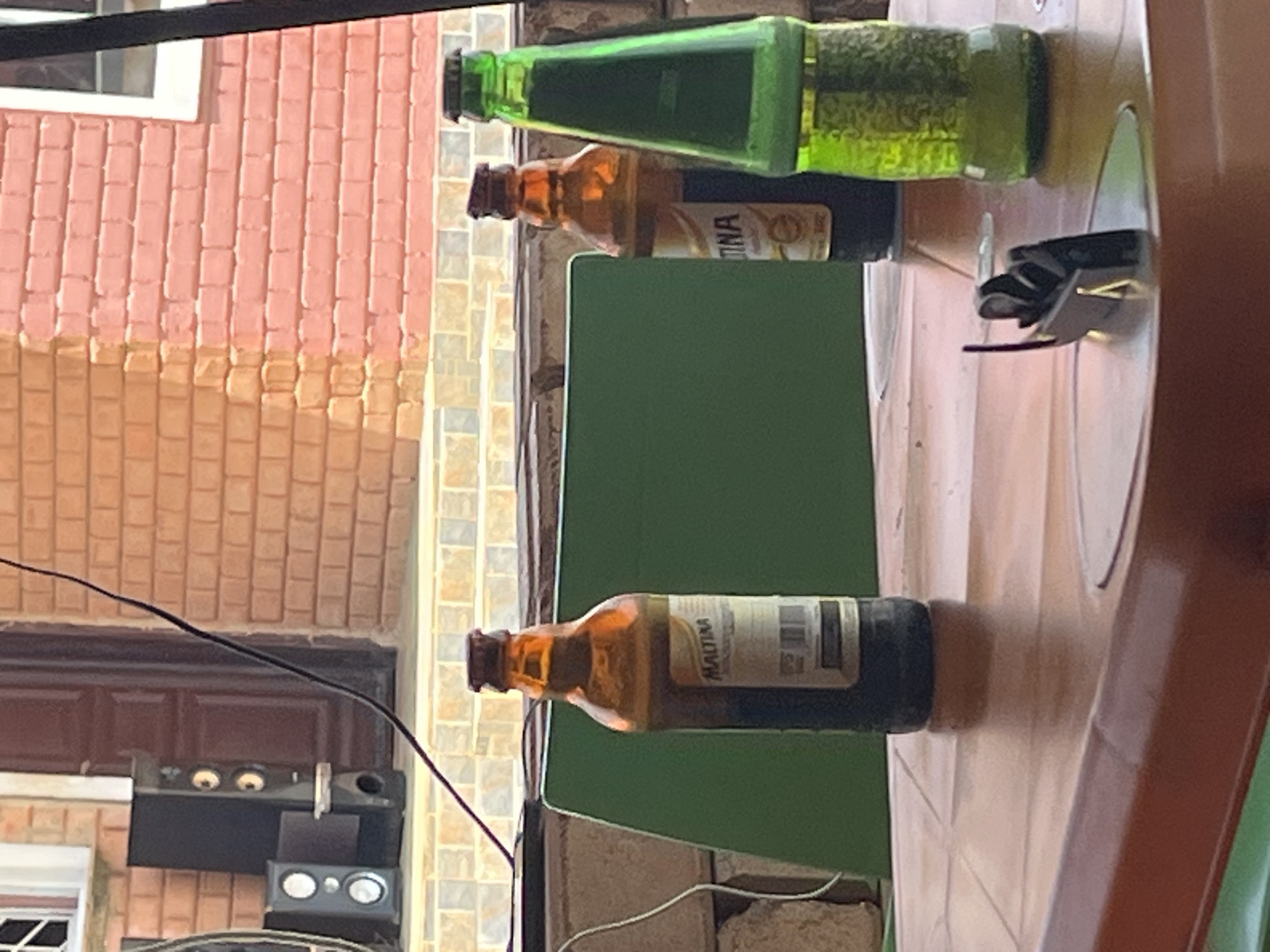 In Jos, Nigeria, on a table, three bottles are displayed. The first bottle on the left is a Maltina bottle, filled with a dark liquid and closed with a brown cap. The middle bottle is also a Maltina bottle, with a dark liquid and a brown cap. The third bottle, on the right, is a green glass bottle filled with a light-colored liquid and capped with a green cap. The table is brown and appears to be made of plastic. Behind the table, a green board is visible, and further back, a brick wall with orange and pink bricks forms the background. Two black speakers are also visible in the background, to the left of the brick wall. A black cable is draped across the scene, originating from the top left and extending diagonally towards the bottom right. The lighting suggests an outdoor setting, possibly during daylight.