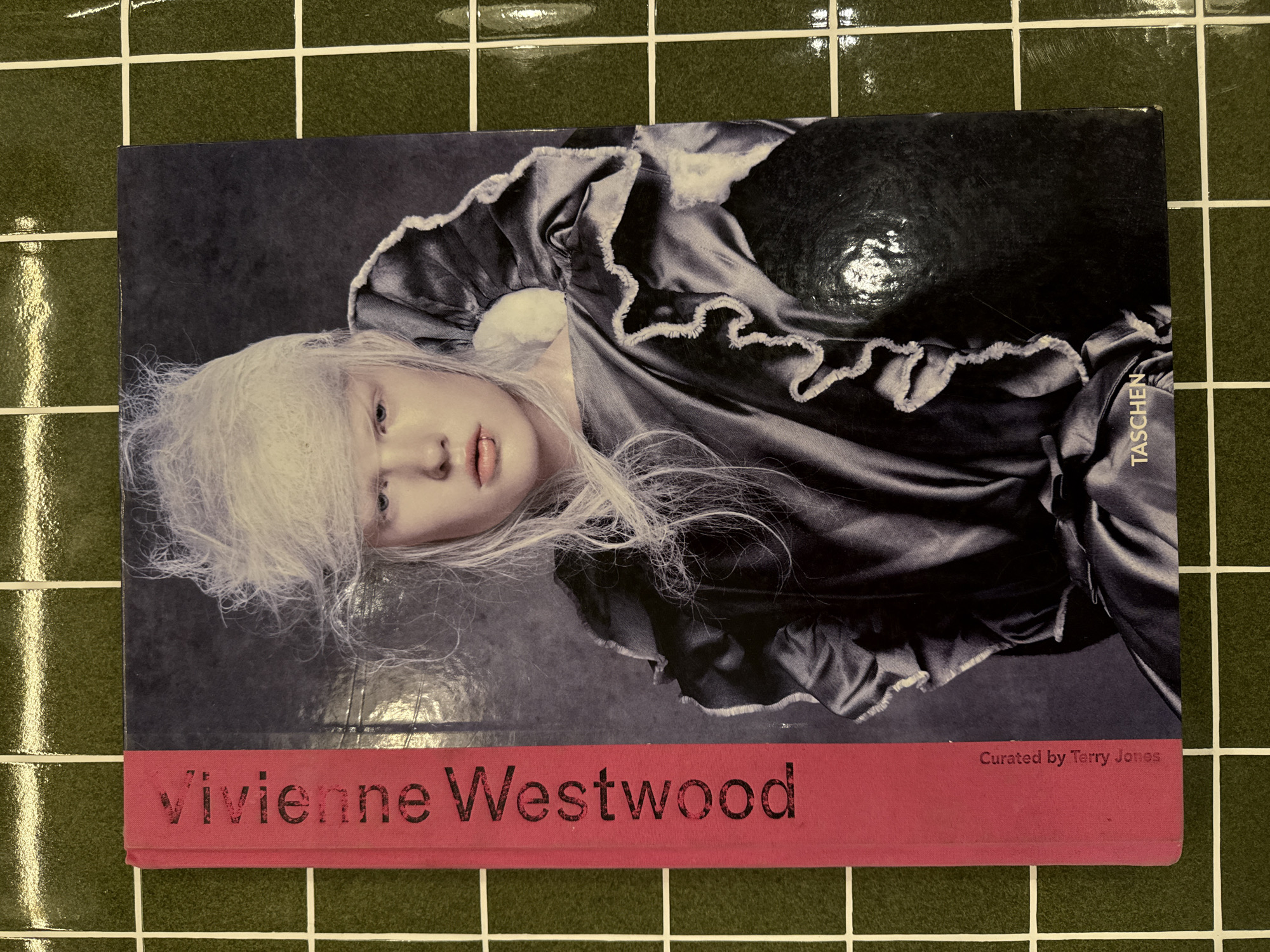 The media file displays a book titled "Vivienne Westwood" lying on a dark green tiled surface. The book's cover features a striking portrait of a person with very pale skin, light eyes, and long, wispy white hair. They are depicted wearing a dark, possibly satin or silk, garment with elaborate ruffles along the neckline and shoulders. The cover image is presented in muted tones, possibly grayscale, highlighting the dramatic contrast with the person's features.
The book is positioned with its spine to the right side of the frame, and the cover image is rotated 90 degrees clockwise, so the subject's head is near the bottom edge of the book. The background consists of square, glossy dark green tiles with prominent white grout lines, forming a grid pattern.
There is no discernible activity taking place; the image is a static shot of the book. Notable details include the glossy finish of both the book cover and the tiles, which show reflections of an overhead light source, particularly in the upper left and upper right sections of the image. The aesthetic of the book cover suggests a publication related to fashion or art.
Visible text on the book cover includes:
- "Vivienne Westwood" in large black letters on a red band across the bottom.
- "Curated by Terry Jones" in smaller white text on the right side of the red band.
- "TASCHEN" in small white vertical text on the right edge of the main cover image, suggesting the publisher.