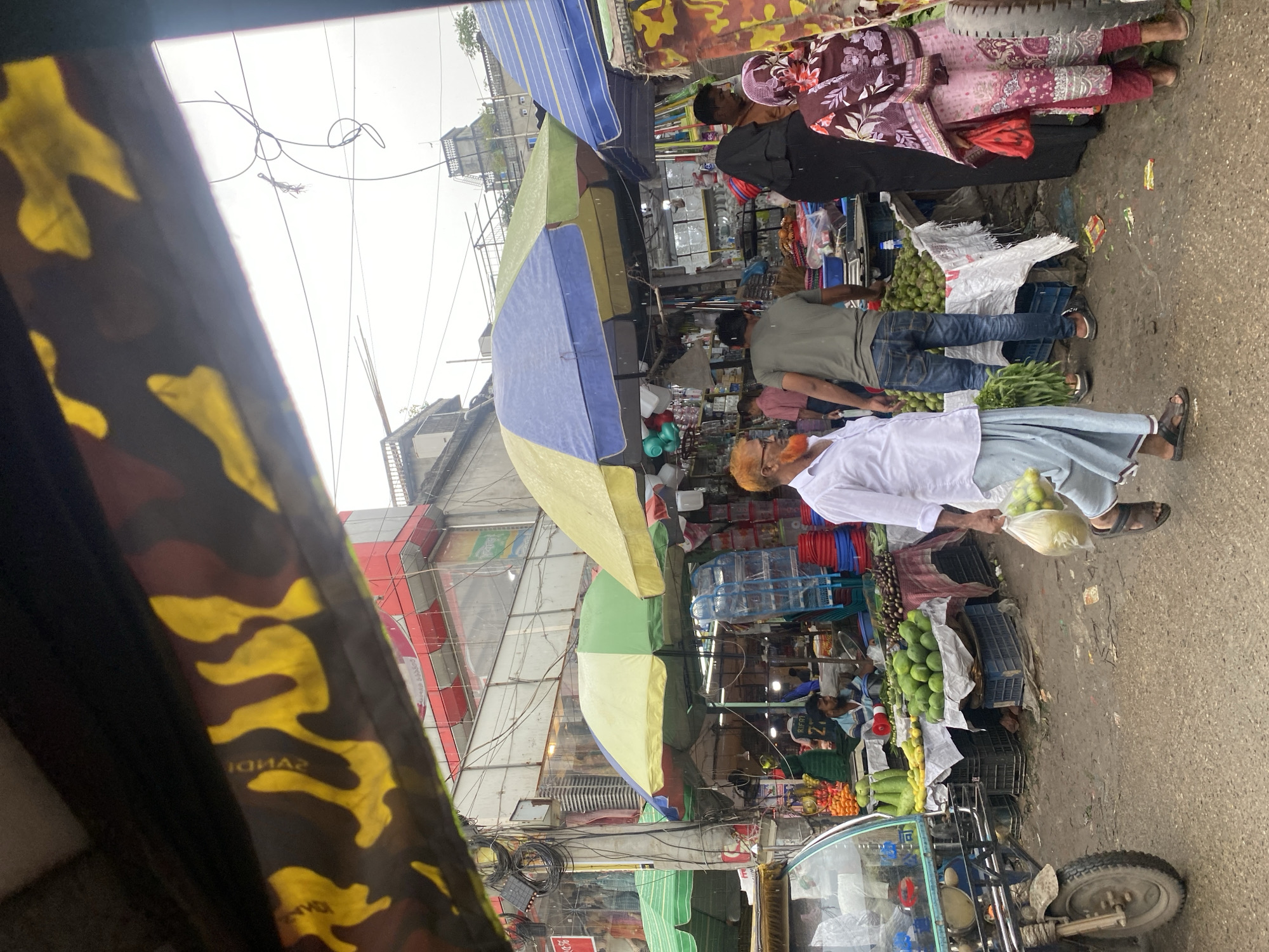 This is a street-level shot of a busy market in Dhaka, Bangladesh, taken from inside a moving vehicle, possibly a rickshaw, as indicated by the camouflage fabric obscuring the top part of the frame. The scene depicts a vibrant marketplace with numerous stalls and vendors. In the foreground, a man wearing a white shirt and a light blue lungi walks carrying two plastic bags filled with green produce. He is wearing sandals and a mask. To his left, a tricycle-like vehicle with a blue canopy is parked.
Further into the market, several stalls are visible, covered by colorful umbrellas, one with yellow and blue panels, another green and yellow, and one to the left with green fabric. These stalls display a variety of goods, including an abundance of green fruits and vegetables, such as limes and what appear to be gourds, arranged on white cloths. There are also piles of eggplants and other produce. Several people are seen tending to the stalls or browsing. One vendor, wearing a green t-shirt and jeans, is attending to his produce. Another individual, wearing a black abaya and headscarf, and a woman in a patterned pink and maroon dress are also present. In the background, buildings with multiple floors are visible, with exposed electrical wires and utility poles adding to the urban landscape. The ground is wet and appears to be unpaved, suggesting recent rain or natural moisture. The overall atmosphere is one of a bustling, everyday market scene in Dhaka.