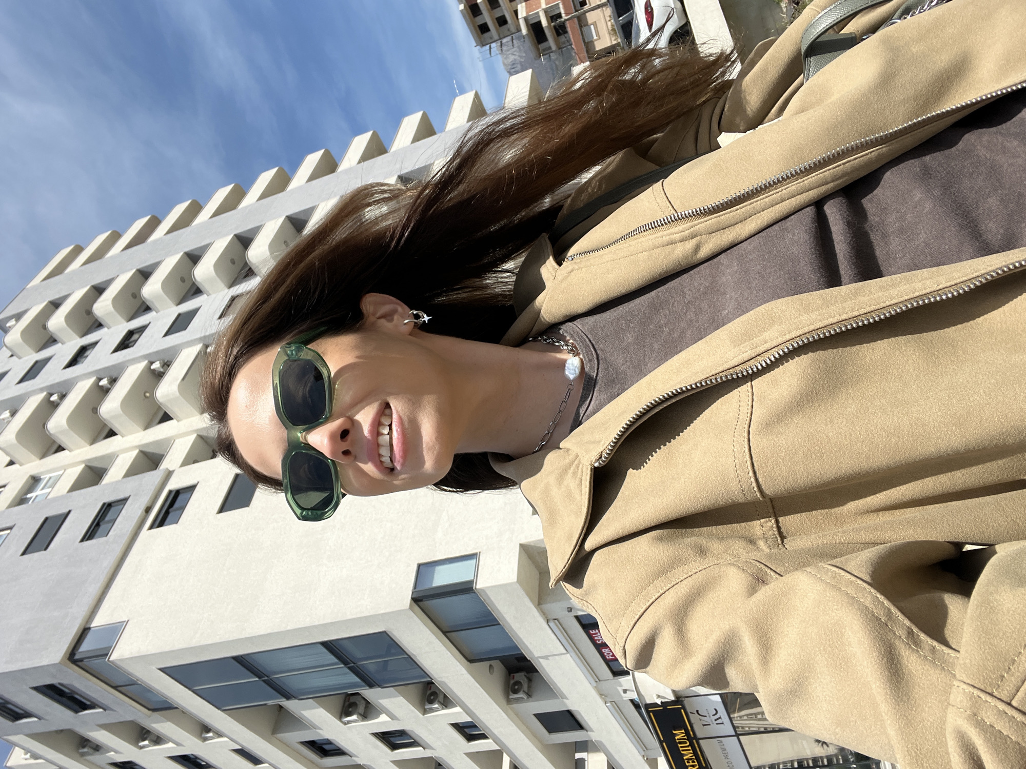 A woman poses outdoors, in front of a modern building. She is wearing green sunglasses, a brown top, and a beige jacket. She also has a silver necklace and an earring. The building behind her has a white facade with windows and protruding balconies. A blue sky is visible above. At the bottom right corner, there's a sign that reads "PREMIUM" and "FOR SALE". The scene is in Bar, Montenegro.