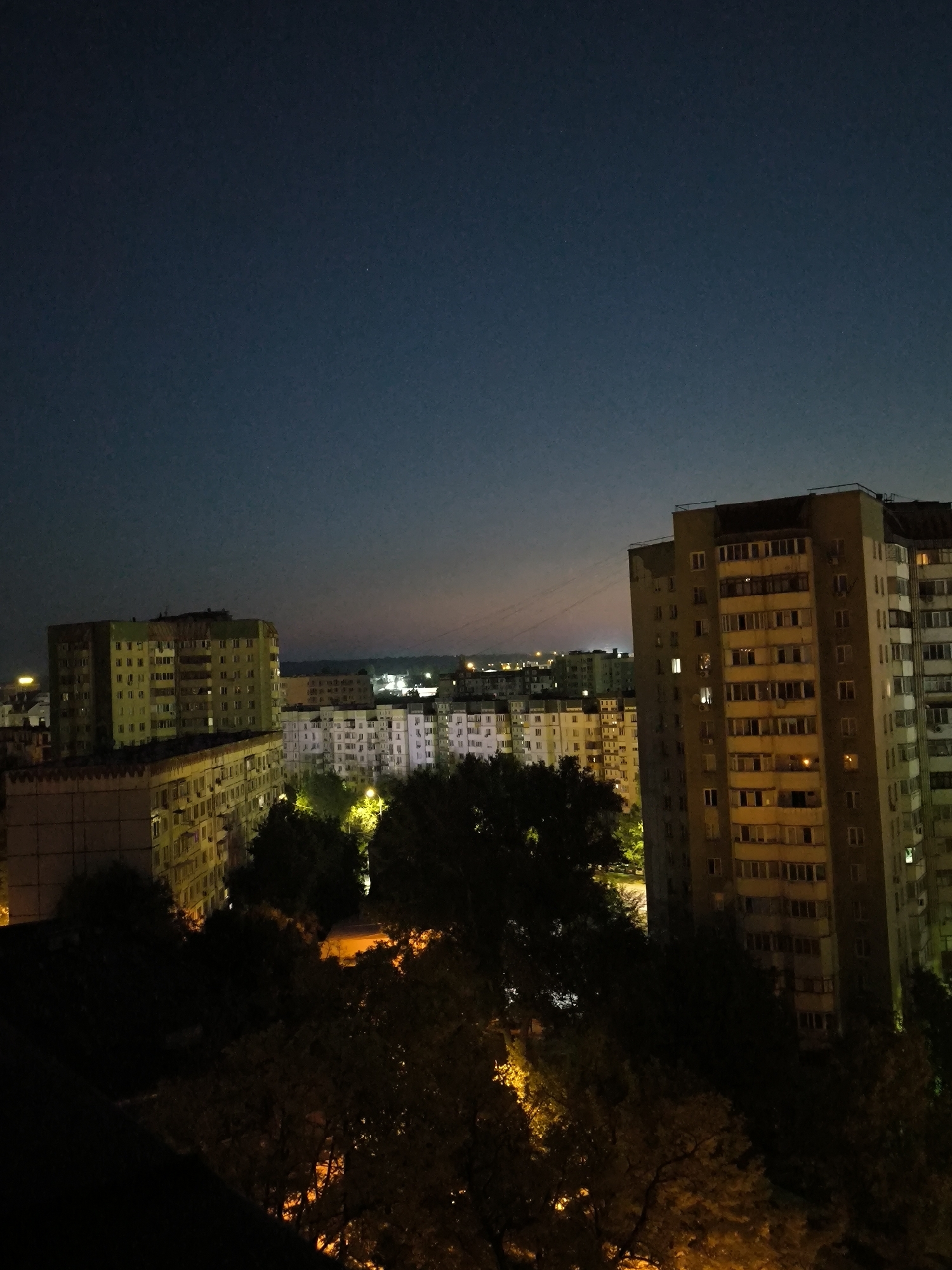 The image captures a nighttime scene of a residential area in Chișinău, Moldova. The sky is dark blue, transitioning to a lighter hue near the horizon, with a few faint stars visible. In the foreground, silhouetted trees obscure parts of the view. Beyond the trees, several apartment buildings of varying heights and architectural styles are illuminated by artificial lights. Some buildings display a warm, yellow glow from interior lights, while others have the cooler white light of streetlamps or exterior lighting. The buildings are tightly packed, suggesting a dense urban environment. The architecture appears to be typical of Soviet-era apartment blocks, characterized by their multi-story design and rows of windows. In the distance, more buildings and lights are visible, hinting at the extent of the city. The overall impression is of a quiet, everyday night in a residential part of Chișinău.