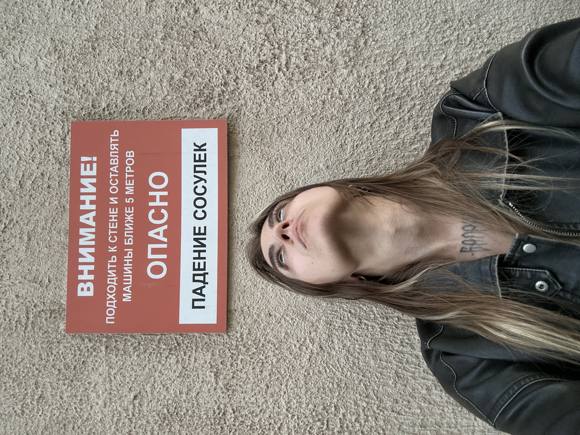 A young woman with long, wavy brown hair, wearing a black leather jacket, looks up at a sign that reads "ВНИМАНИЕ! ПОДХОДИТЬ К СТЕНЕ И ОСТАВЛЯТЬ МАШИНЫ БЛИЖЕ 5 МЕТРОВ. ОПАСНО. ПАДЕНИЕ СОСУЛЕК" in Russian, which translates to "ATTENTION! APPROACH THE WALL AND LEAVE CARS WITHIN 5 METERS. DANGEROUS. FALLING ICE." The sign is mounted on a textured concrete wall. The woman has distinctive makeup on her eyes, with dark eyeliner and visible eyelashes. A tattoo is visible on her chest. The background is an exterior wall, suggesting an outdoor urban environment, likely in Moscow, Russia, due to the Russian language on the sign and the architectural style of the wall. The lighting suggests it is daytime.