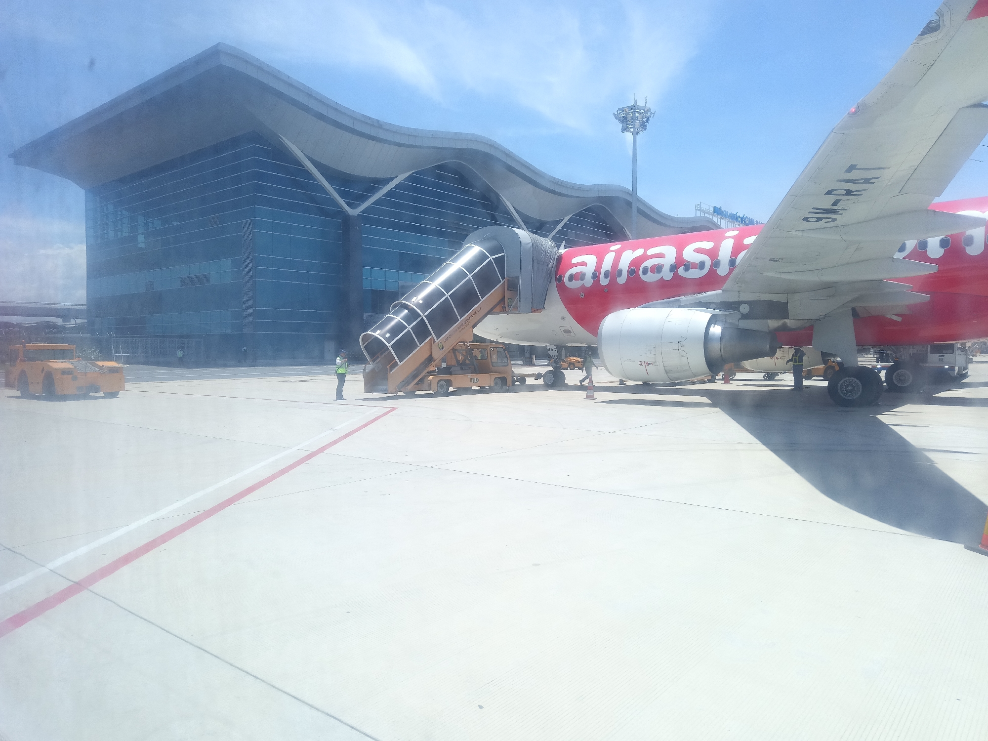 This image depicts an airport tarmac at Cam Ranh City, Vietnam. A red AirAsia airplane is parked, with a mobile staircase attached to its side. The plane's registration number, 9M-RAT, is visible on its wing. Ground crew members are present near the aircraft, attending to operations. In the background, a modern airport terminal with a distinctive wave-like roof design is visible. Yellow airport vehicles, likely baggage tugs or service carts, are positioned on the tarmac. The scene is set under a clear blue sky, suggesting daytime.
