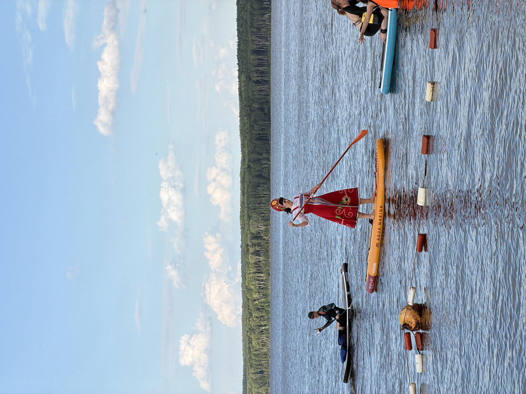 Two people are paddleboarding on a calm, blue lake. In the foreground, a person in traditional Russian attire, including a red and white embroidered dress and a red headscarf, stands on an orange paddleboard, holding a paddle. In the mid-ground, a second person is kneeling on a black paddleboard, wearing a dark jacket and a cap. They are both looking towards the viewer. The lake is surrounded by a dense, green forest. The sky is a clear blue with scattered white clouds, suggesting it is a sunny day, likely in the afternoon. Several buoys are visible in the water, marking a course or area. The overall scene depicts a recreational activity on a body of water with a backdrop of nature. The person in traditional clothing may be participating in a cultural event or simply enjoying a themed outing. There is text visible on the orange paddleboard: "AQUA MARINA" and "FUSION" along with "10'6\"x32\"". The location is identified as Chaikovsky, Russia.