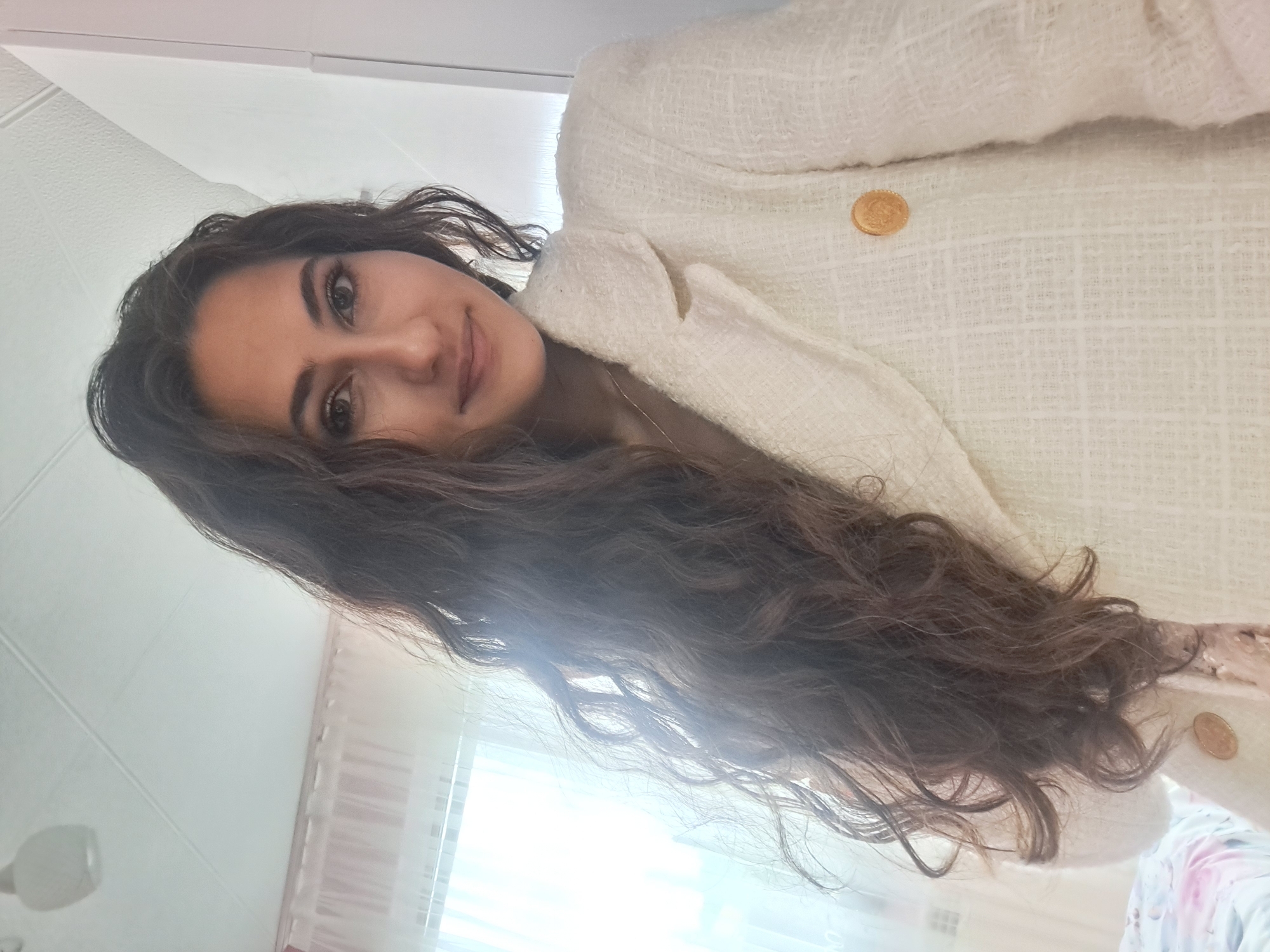 A young woman with long, dark, wavy hair is pictured from the chest up, looking into the camera. She is wearing a white, textured jacket with gold buttons. The background is a softly lit room with sheer curtains visible on the left, suggesting a window is present. The ceiling with a light fixture is also visible at the top of the image. The image appears to be a selfie taken indoors.