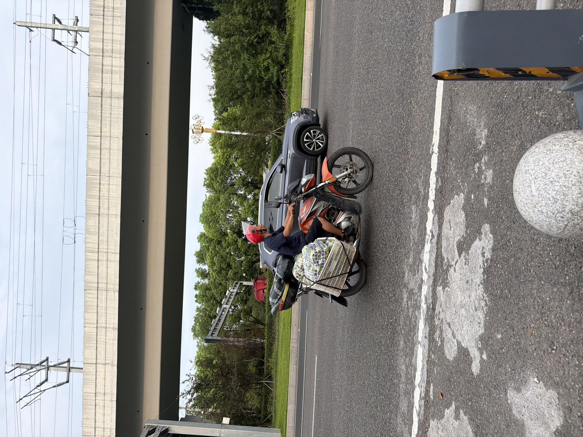 In the city of Jiangjin, China, a man wearing a red helmet rides an orange motorcycle laden with cargo. He is on a paved road with a white line marking the lane. In the background, a grey SUV is driving in the adjacent lane. Above the road, a concrete overpass stretches across the scene, supported by pillars. Overhead, power lines are strung between utility poles. The environment is largely urban with lush green trees lining the roadside. The man is carrying a large woven basket filled with produce, possibly watermelons due to their green and white striped pattern. Other items, wrapped in black plastic bags, are secured to the rear of the motorcycle. The overall lighting suggests it is daytime.