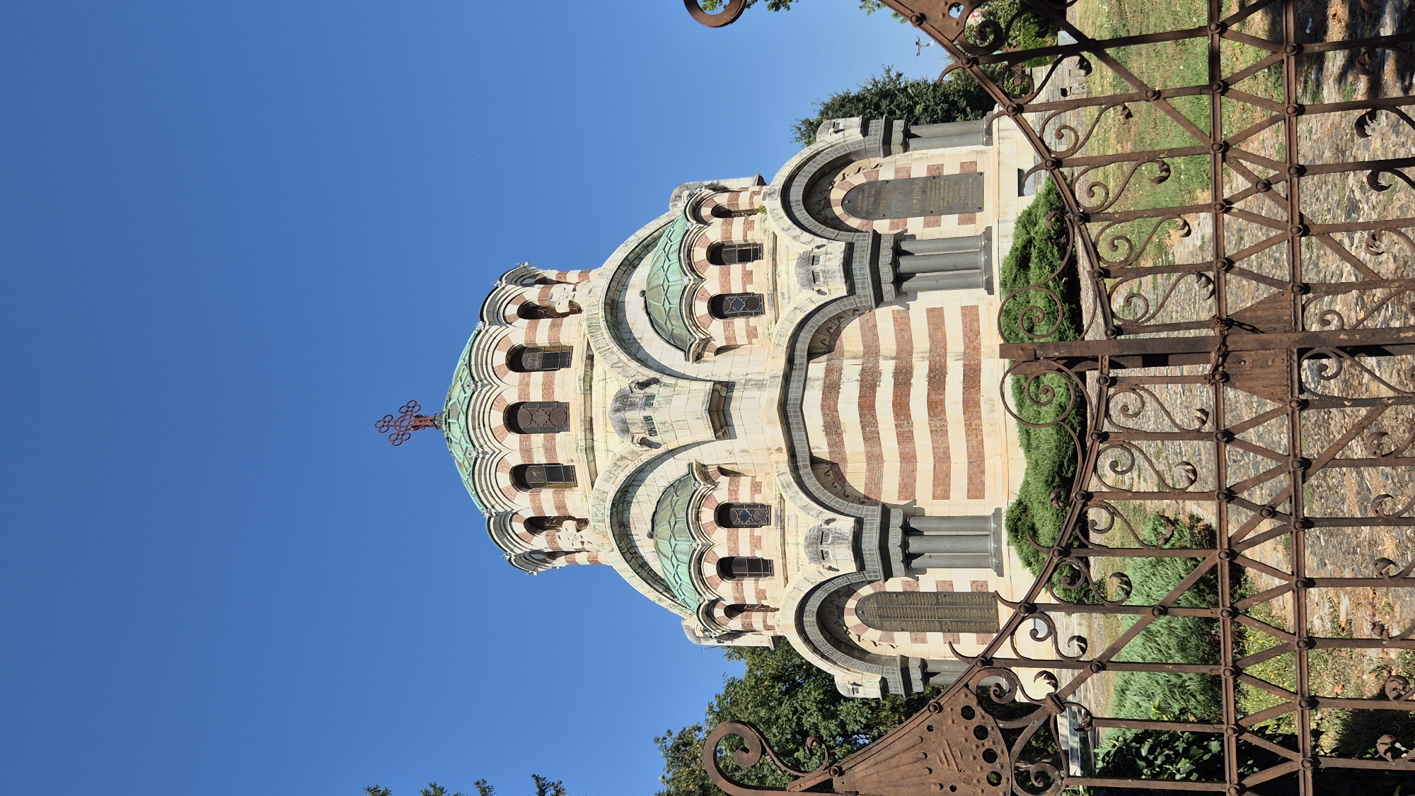 The image shows the Pleven Mausoleum, also known as the Chapel-Mausoleum of the Liberation of Pleven, located in the city of Pleven, Bulgaria. The mausoleum is a striking structure with a central dome topped by a red Orthodox cross, surrounded by smaller domes. The building is constructed with alternating red and white horizontal bands, with arched windows and columns. In the foreground, a rusty, ornate metal gate partially obscures the view of the mausoleum and its surrounding greenery. The sky above is clear and blue, indicating a sunny day. The grounds appear to be a park or memorial area, with manicured bushes and paved pathways visible behind the gate.