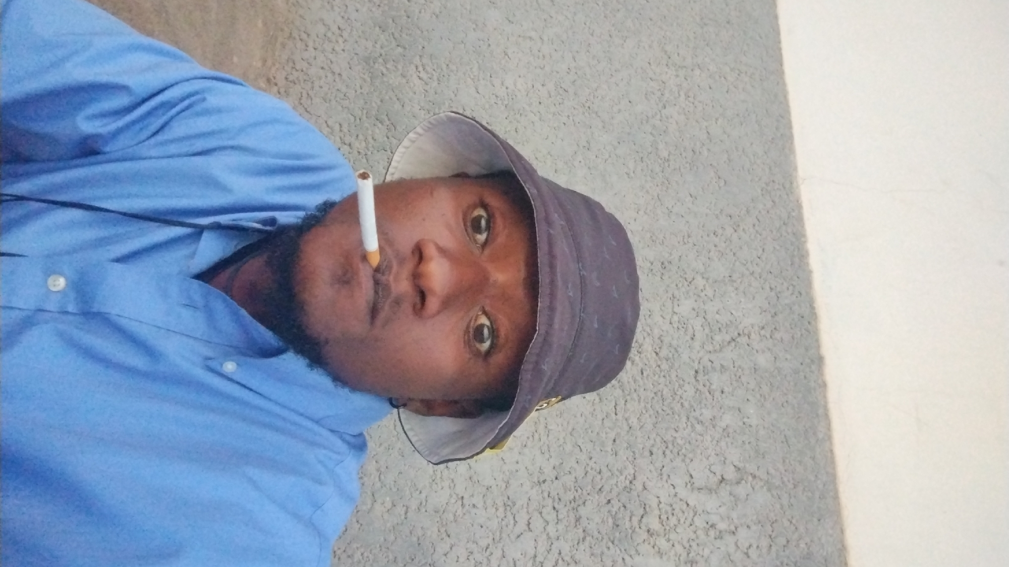 A man with dark skin, a beard, and wearing a blue shirt and a bucket hat with a gold or yellow brim is looking directly at the camera with a cigarette in his mouth. The background consists of a wall with a textured gray surface on the left and a smooth white or light-colored surface on the right. The location is in Jos, Nigeria.