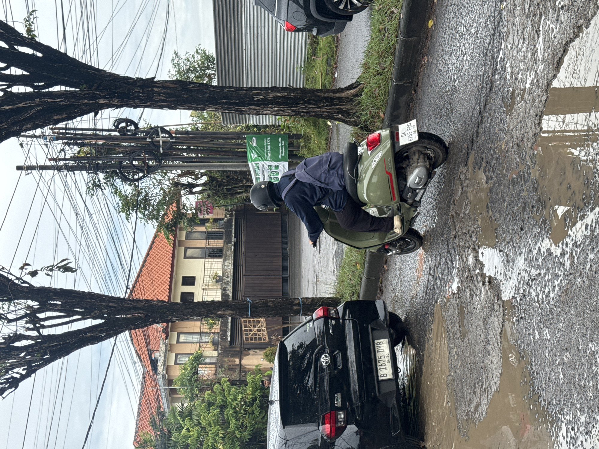 The image captures a street scene in Kuta Selatan, Indonesia, on an overcast and wet day. In the foreground, a person wearing a dark blue jacket and a black helmet is riding a green scooter, resembling a Vespa, through muddy puddles on an uneven road surface. The scooter's rear license plate, "DK 6755 FDG," is visible, indicating Bali registration with an expiry date of June 2029.
To the left, a black Toyota SUV, branded with "ASTRA," is parked partly in a large puddle. Its license plate reads "B 1675 DYB," suggesting Jakarta registration with an expiry of April 2030. The street is lined with large trees featuring dark, textured trunks, with one tree extending horizontally across the upper frame.
Above the street, a dense array of utility wires is tangled between poles. In the background, buildings with distinctive red-tiled roofs and multiple windows are visible. A green banner attached to a pole displays "NT DUURBAN," a phone number "0812-388-1771," and an address "Jl. Diponegoro No. 222 Denpasar - Bali," firmly placing the location within Bali. The presence of numerous puddles and mud indicates recent rain.