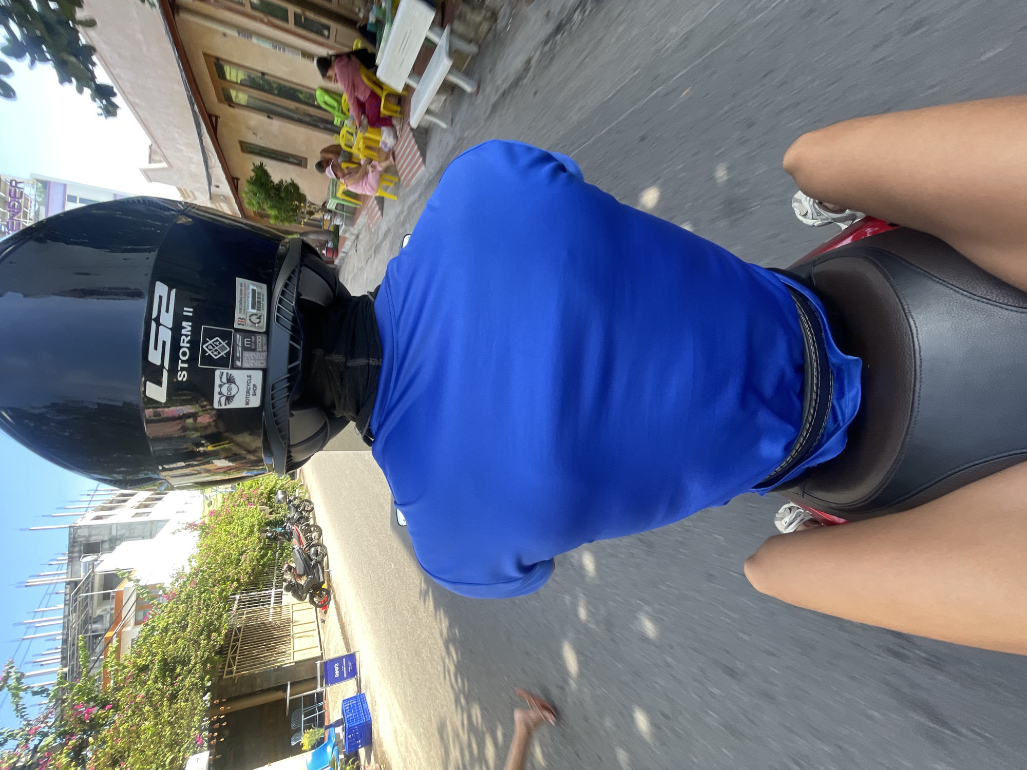 The image shows a person riding a motorcycle in Mũi Né, Vietnam. The rider is wearing a blue shirt and a black helmet. The motorcycle is on a paved road, with buildings and trees on the side. There are several other motorcycles parked on the side of the road, and some people are sitting at tables outside a building. The sky is clear and blue.