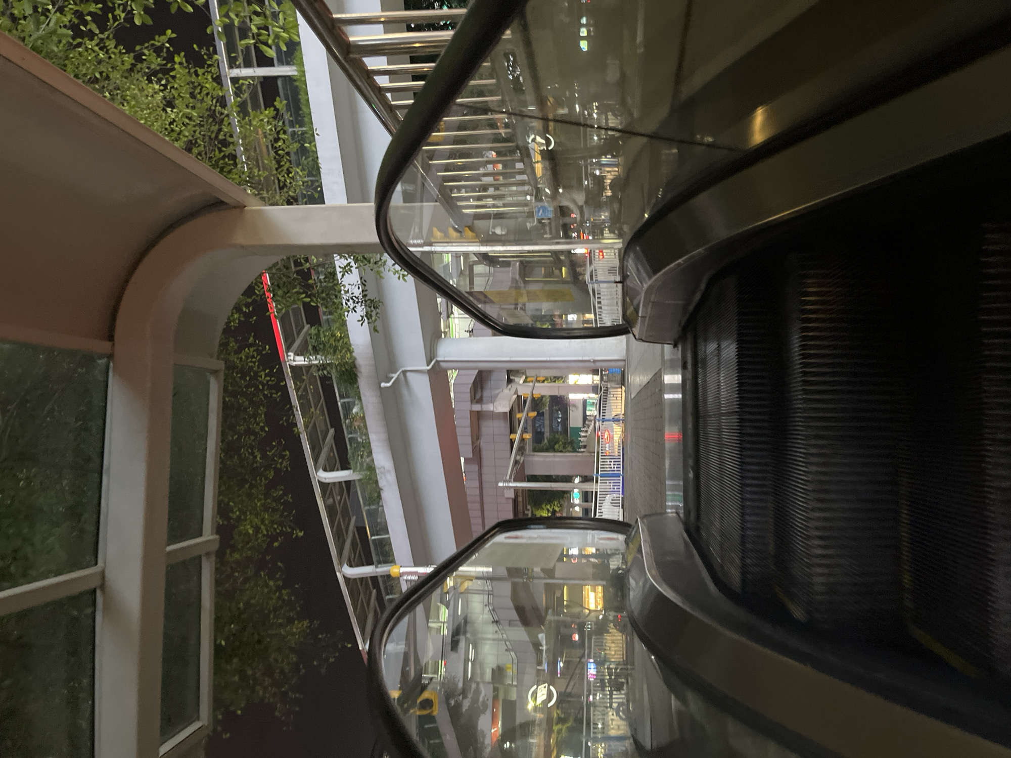 The image shows a view from the bottom of an outdoor escalator in Jiulongpo District, China. The escalator is moving upwards. The scene appears to be at night, as indicated by the artificial lighting and dark sky. The escalator has glass panels on either side, reflecting the surrounding environment. In the background, there are elevated walkways and buildings, with greenery visible on some structures. Traffic lights and streetlights are also present. There are no people clearly visible on the escalator or in the immediate foreground. The overall environment suggests an urban setting, possibly a transportation hub or a pedestrian overpass area.