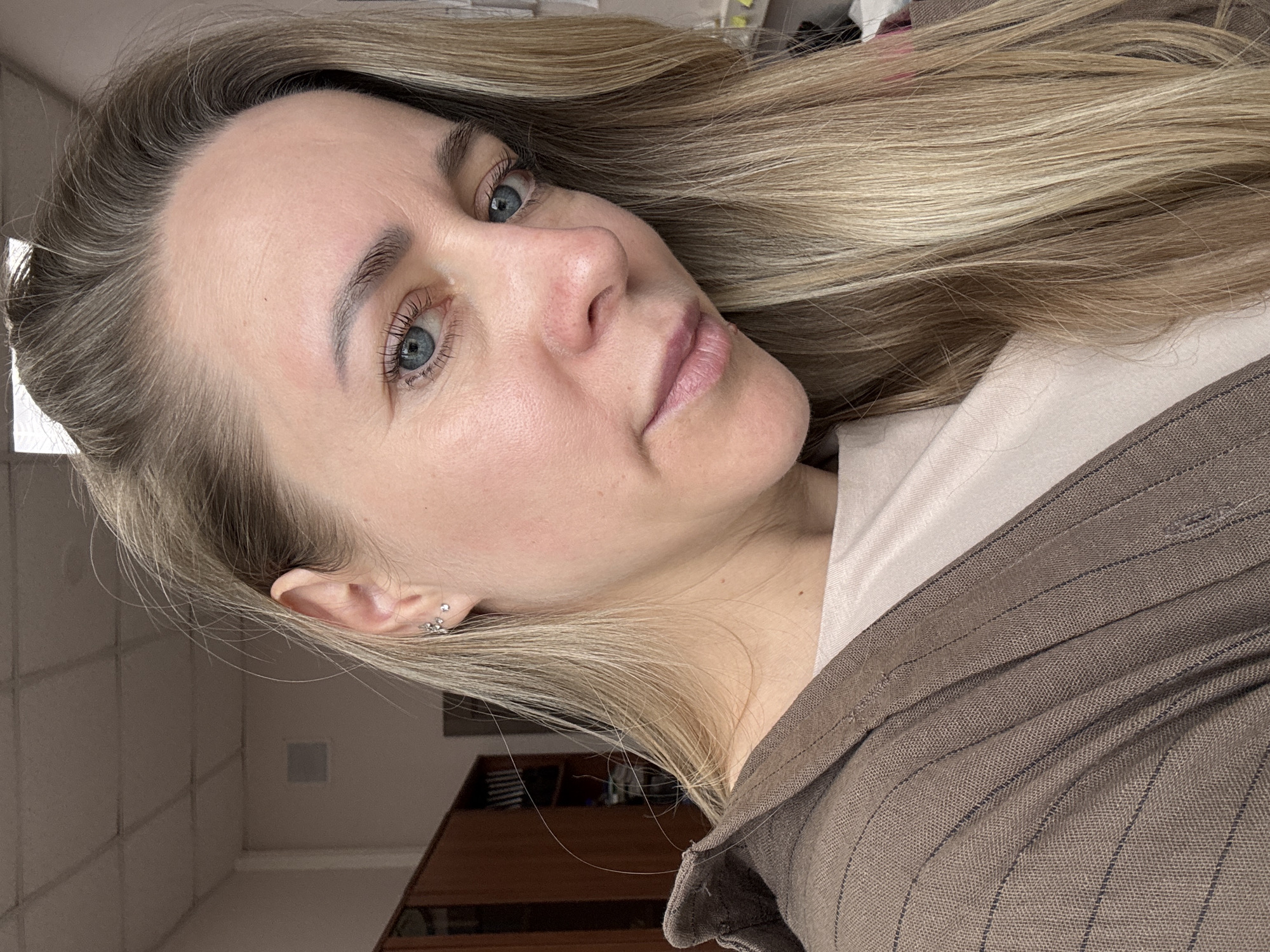 A close-up portrait of a fair-skinned woman with light blonde hair and blue eyes. She is facing the camera at an angle. She has light eyebrows and eyelashes. She is wearing a beige top under a brown pinstripe blazer. Her left ear is visible, with multiple earrings. The background is blurry, showing a light-colored wall and a piece of furniture. No information to determine this is in Chaikovsky, Russia.