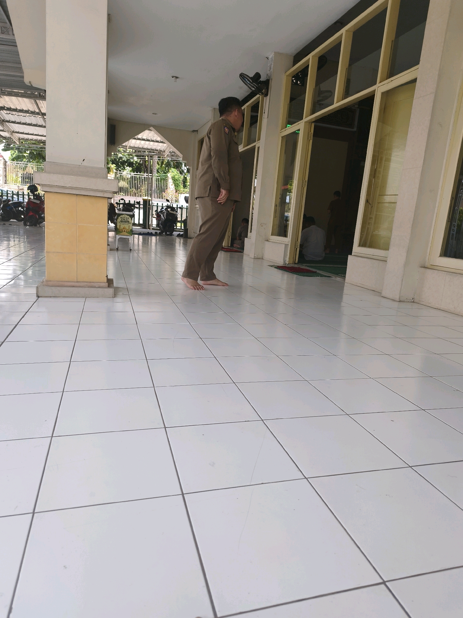 The image captures a street scene in Samarinda, Indonesia, with a focus on the exterior of a building that appears to be a mosque or a similar religious institution. The foreground is dominated by a tiled walkway, likely an outdoor prayer area or verandah, with large, light-colored square tiles.
In the mid-ground, a man in a brown uniform, possibly a security guard or government official, is seen walking barefoot. He is turned away from the camera, facing towards the building. Behind him, several motorcycles are parked, suggesting this is an area where people arrive by vehicle. Further back, through an open doorway or window, other individuals can be seen inside the building, some of whom are in prayer. The building itself has a modern aesthetic with large glass-paned windows framed in yellow. A ceiling fan is visible mounted on the exterior wall of the building. The overall lighting suggests it is daytime, possibly overcast or in the shade. The presence of parked motorcycles and the context of individuals praying inside indicate a functional religious or community space.