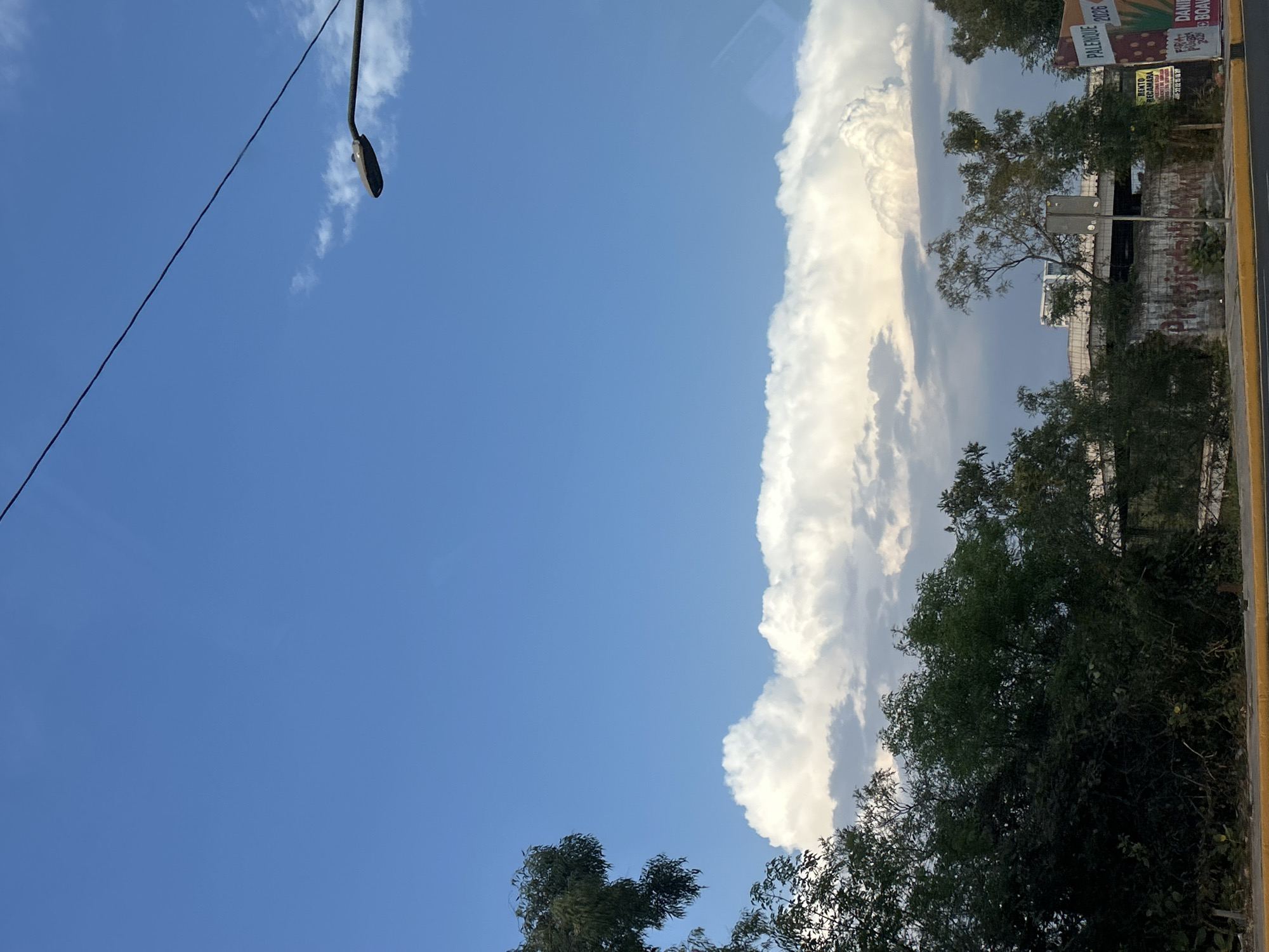 The photograph captures a daytime scene in Puebla, Mexico, as suggested by the presence of signage written in Spanish. A clear blue sky dominates the left portion of the frame, contrasting with a tall, billowing cumulus cloud formation situated on the right.
In the upper-left corner, a street lamp and its supporting electrical wires are visible. The lower section of the image features treetops, partially obscuring a view of a building and a wall covered in graffiti. A yellow roadside barrier runs along the far right edge of the frame, bordering the street. The scene lacks any discernible human presence or activity.