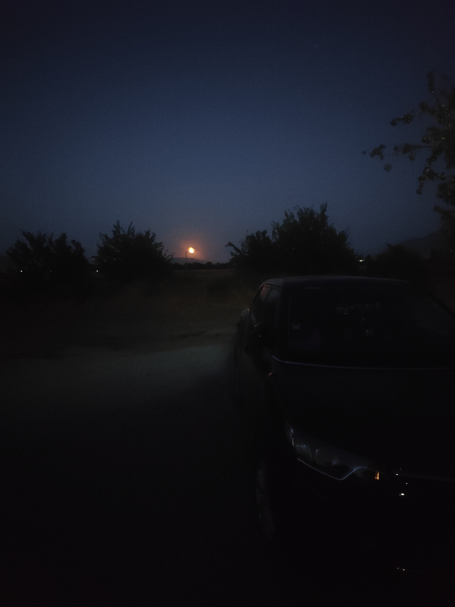 The image depicts a dark, rural scene with a car in the foreground, partially obscuring the view of the landscape. The sky is a deep twilight blue, with a faint, orange-hued object, possibly the moon or a distant light source, visible low on the horizon. Silhouetted trees and shrubs form a dark line across the middle ground. A dirt road or path leads away from the car into the darkness. The overall atmosphere is still and quiet, suggesting a rural or semi-rural setting outside of an urban center. The location is stated to be in the city of Байгелди, Kyrgyzstan.