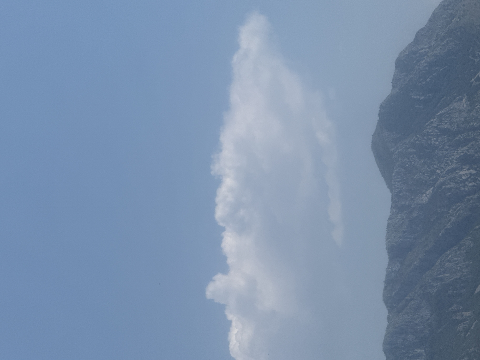 This media file captures a dramatic juxtaposition of a cloudy sky and a rugged mountain face. The scene is dominated by a tall, wispy cloud formation that appears to be a cumulus cloud, rising vertically against a pale blue sky. To the right, a steep, dark gray mountain slope dominates the frame, its textured surface revealing the geological formations of rock and possibly some sparse vegetation. The lighting suggests it is daytime, with the sun likely positioned behind or to the side of the viewer, illuminating the cloud and casting subtle shadows on the mountain. The overall impression is one of natural grandeur and vastness. No people, objects, text, or specific activities are visible. The location context of Boreti, Montenegro, suggests this could be a view of the Montenegrin coast or inland mountainous region. The weather appears fair with some cloud cover.