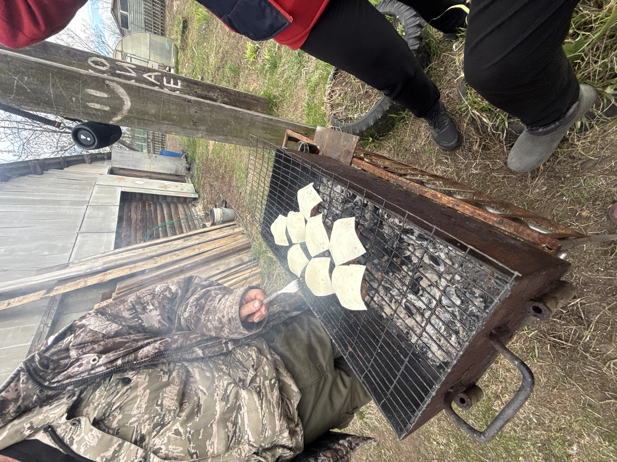 An outdoor scene in what appears to be a rural or backyard setting in Кемуль, Russia, shows people grilling. The image is tilted, giving a dynamic perspective.
In the foreground, a person wearing a camouflage jacket and green pants is tending to a rectangular metal grill (mangal) filled with charcoal and a metal grate. On the grate, several square slices of white cheese are melting on top of what appear to be burger patties, generating smoke. The person is holding a metal spatula, suggesting they are actively cooking.
Two other individuals, or parts of them, are visible standing around the grill. One person is wearing black leggings or pants and dark athletic shoes. Another leg is visible, wearing a grey rubber boot. A person's torso, clad in a red and blue jacket, is seen near the grill.
The setting includes various rustic elements: old, weathered wooden buildings and shacks are visible in the background, along with a small greenhouse with a rounded roof. The ground is a mix of sparse green grass and dirt. An old car tire is partially buried in the ground nearby.
A large wooden beam or post runs vertically through the left side of the frame. Attached to it is a black cylindrical JBL portable speaker. White text is visible graffitied on the wooden beam, spelling "МАШЕ" (MASHE), along with a simple smiley face drawn next to it. The sky appears overcast, indicating a daytime setting without direct sunlight. The overall atmosphere suggests a casual, outdoor cooking event.