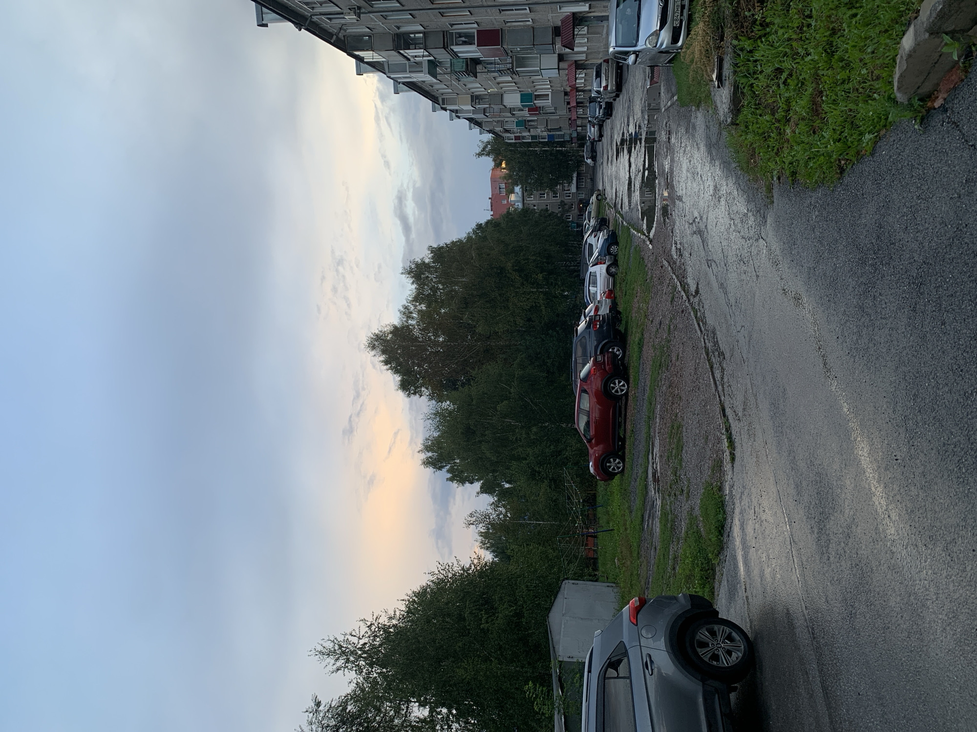 The image depicts a residential street in Prokopyevsk, Russia, after a rainfall. The scene is dominated by a row of multi-story apartment buildings on the right, with balconies and weathered facades. On the left, a dense line of trees separates the street from what appears to be a parking area and a small, grey shed. The asphalt road is wet and reflects the overcast sky, with several large puddles visible, indicating recent precipitation. A variety of parked cars are present, including a silver SUV in the foreground on the left, a red hatchback further down the road, and other vehicles parked along the street and in the designated area. The sky is a blend of soft blues and greys, with hints of pale orange near the horizon, suggesting either dawn or dusk. There are no people visible in the frame. The overall impression is a quiet, everyday urban scene in a post-rain setting.