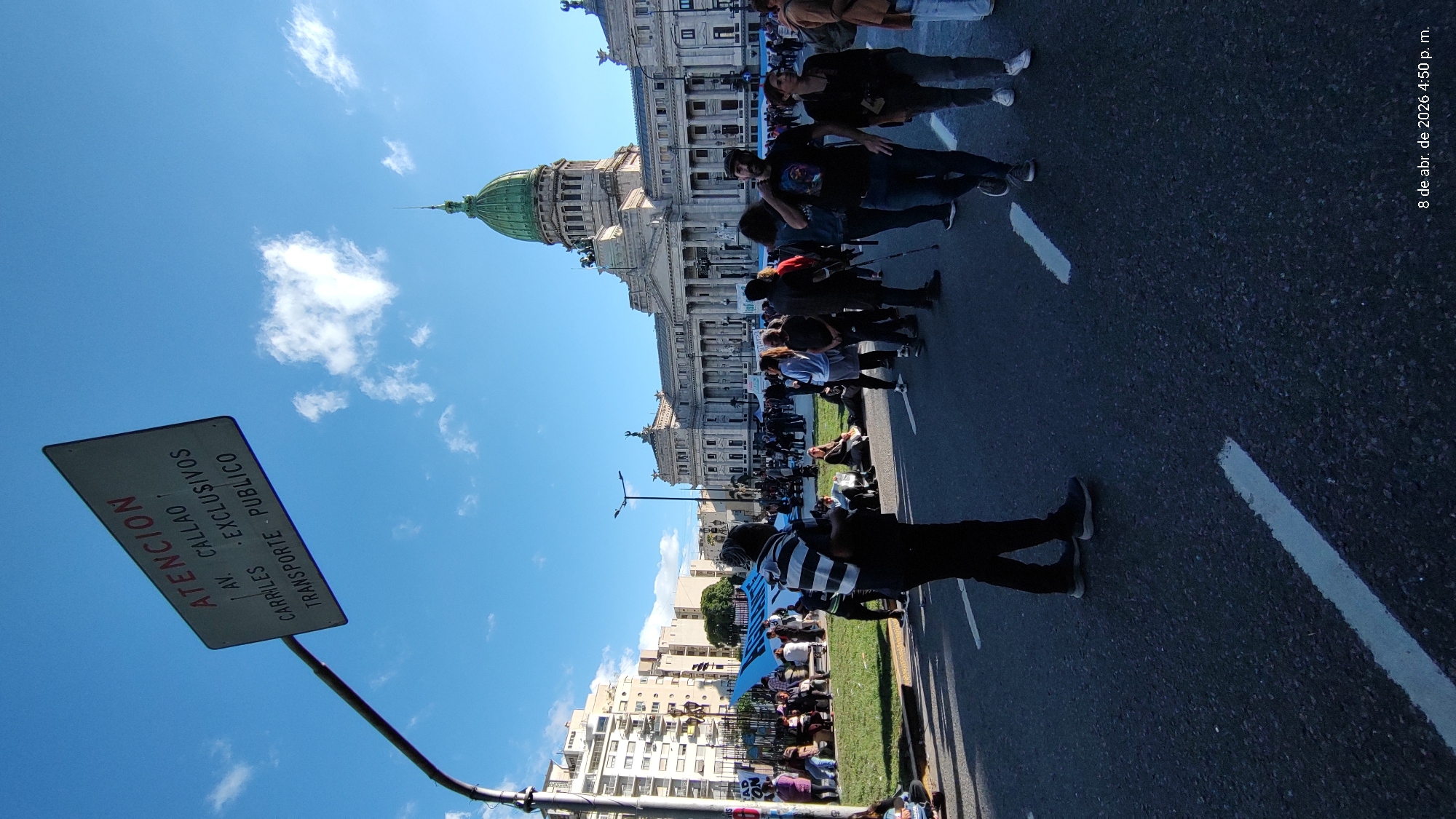 The image depicts a street scene in Buenos Aires, Argentina, with a crowd of people gathered on the pavement and road. In the background is a large, ornate building with a green dome. The dome suggests the building may be the Argentine National Congress. In the foreground is a road sign with the words "ATENCION" "AV. CALLAO" "CARRILES" "EXCLUSIVOS" "TRANSPORTE PUBLICO". The sky is blue with scattered clouds. The time stamp on the image says: 8 de abr. de 2026 4:50 p. m. Several people are standing along the curb, while others are walking along the road and standing on the grass. There are also several residential buildings to the left of the crowd.