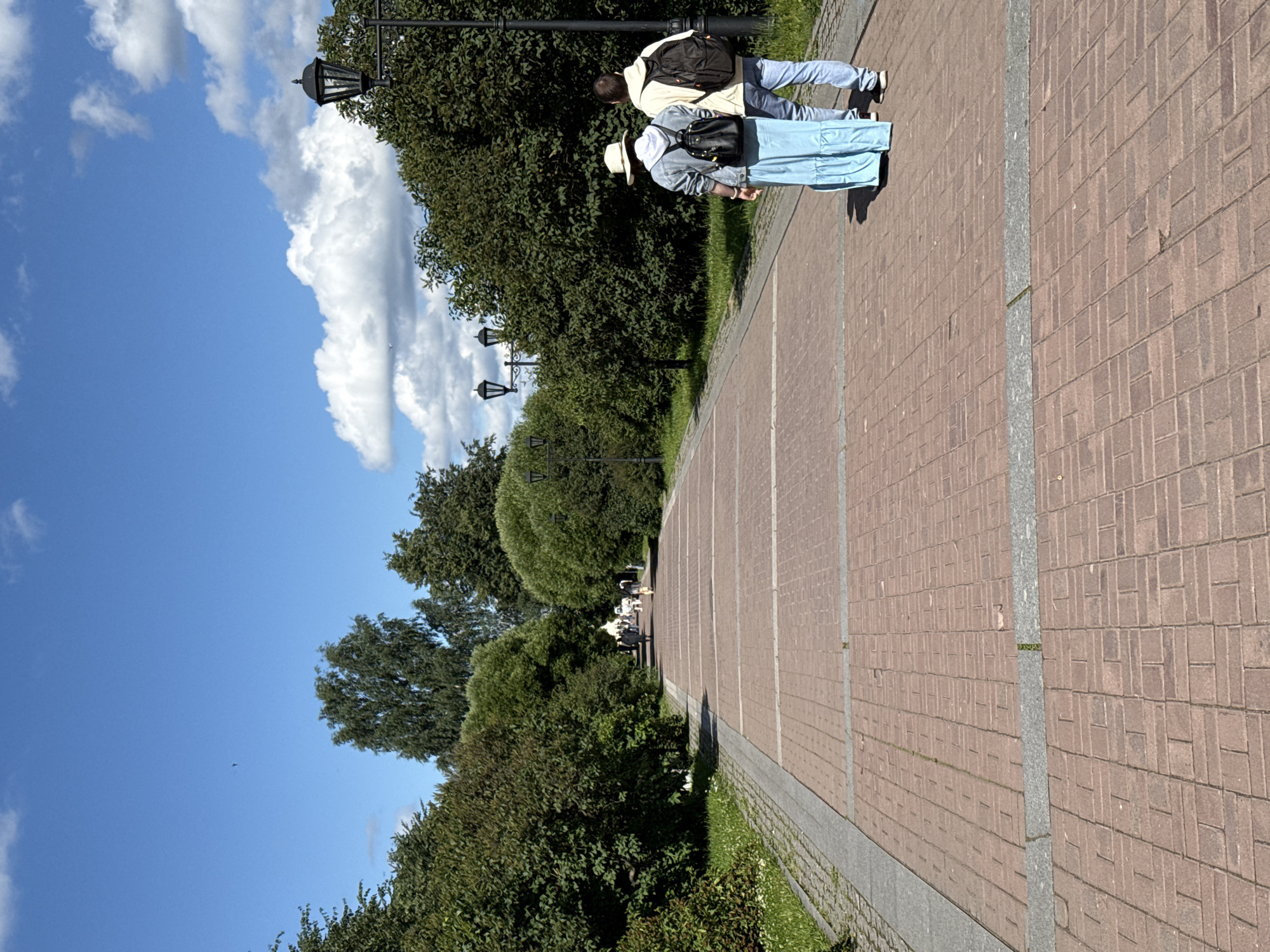 A wide, red brick walkway lined with green trees stretches into the distance under a blue sky with scattered white clouds. Two people, a woman wearing a wide-brimmed hat, denim jacket, and long blue dress, and a man in a yellow hoodie and jeans, walk away from the camera on the right side of the walkway, both carrying backpacks. Further down the walkway, several other figures are visible, appearing to walk or stand amidst the trees and lampposts that line the path. The scene suggests a park or public garden.