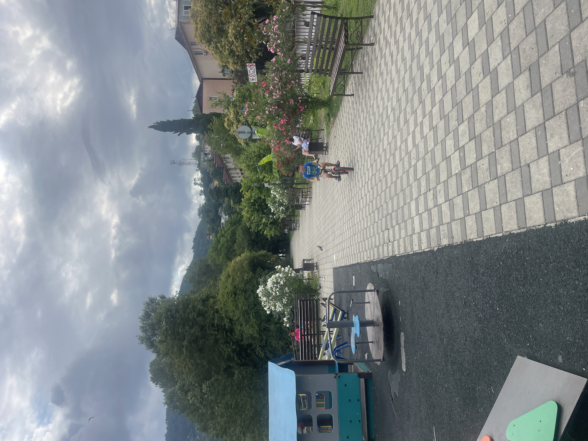 The image depicts a public park area on a cloudy day, likely in Sochi, Russia, given the architecture and vegetation. In the foreground to the left, a children's playground features a blue and gray play structure with a small merry-go-round on a dark rubberized surface. A woman in a bright pink top is seated on a bench behind the playground.
The main area of the image is a wide, paved walkway made of interlocking gray and beige square pavers. A young boy rides a red bicycle along this walkway. Further down the path, several park benches are visible, some occupied by people. One woman is sitting on a bench, reading a book. Lush green trees and flowering bushes, with pink and white blossoms, line the walkway. In the background, buildings are visible on a hillside, interspersed with more trees, suggesting a coastal or mountainous region. A tall, slender cypress tree stands out against the sky. The sky is overcast with dramatic clouds. Pigeons are present on the walkway and grass.