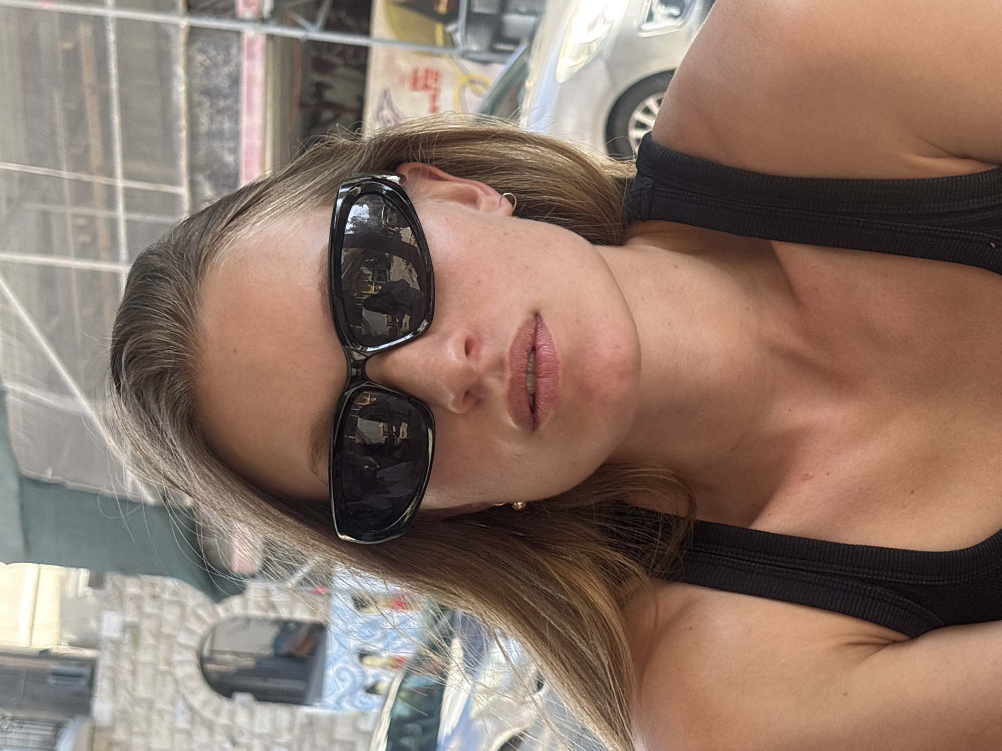 A young adult female is depicted in a head-and-shoulders portrait, looking directly at the camera. She wears a black ribbed tank top and dark, wraparound sunglasses that obscure her eyes. Her long, light brown hair is parted on the side, with some strands visible. Her skin has a tanned complexion, and her lips are slightly parted. She wears small, gold-colored earrings: a hoop in her right earlobe and a spherical stud in her left. The reflections in her sunglasses show indistinct indoor elements, including multiple figures, suggesting she is situated in an establishment such as a cafe or restaurant.
The background establishes an urban setting in Tel-Aviv, Israel. To the left, a light-colored stone building with an arched opening and a blue and white patterned wall is partially visible. A silver or white car is also seen behind her left shoulder. Above the building, a green tarp or awning is present. To the right, extensive metal scaffolding indicates ongoing construction or renovation, with another silver or white car visible behind her right shoulder. The scene is illuminated by bright daylight.