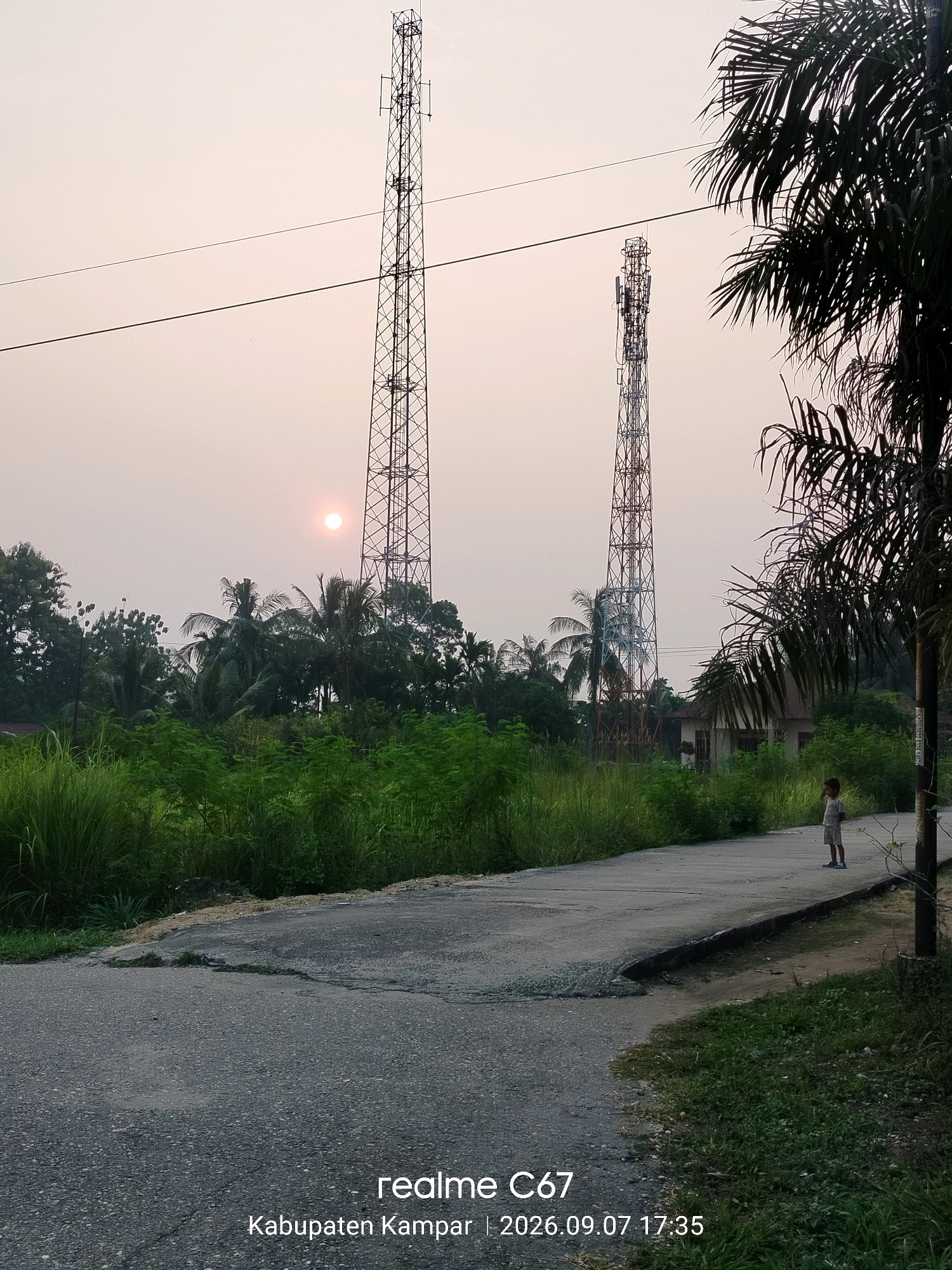A young child stands on the side of a paved road in Pandau Jaya, Indonesia. The child is facing away from the camera, looking towards two large telecommunication towers in the distance. The towers are tall, lattice structures, one appearing taller and more slender than the other. Between the towers and the road, there is dense green vegetation, including tall grasses and trees, with palm fronds visible in the foreground to the right. The sun is visible low in the sky, casting a warm glow over the scene, suggesting it is either sunrise or sunset. The road has a rough, cracked surface. A house is partially visible behind the vegetation and to the left of the smaller tower. The image has a watermark at the bottom stating "realme C67 | Kabupaten Kampar | 2026.09.07 17:35".