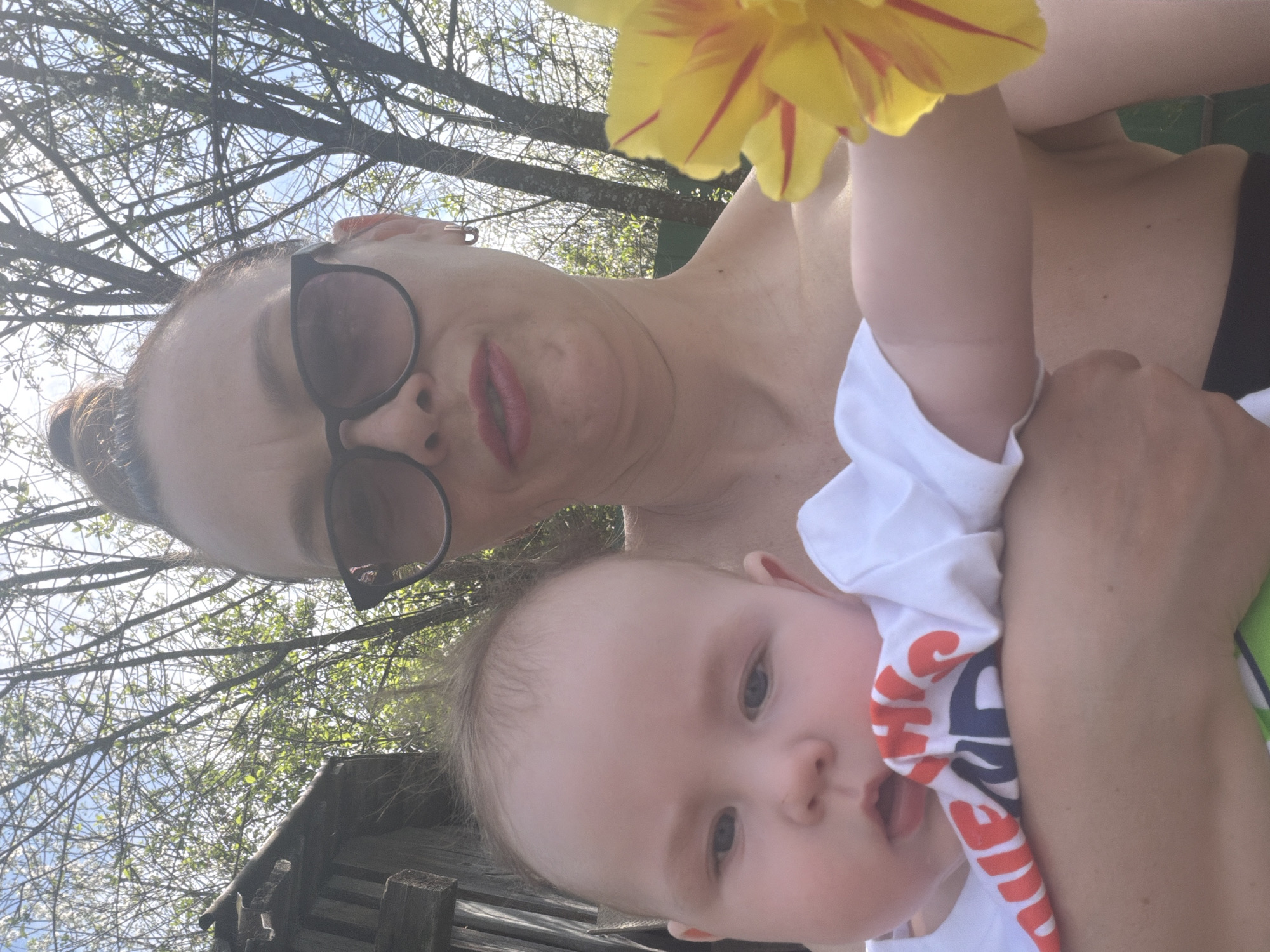 A medium shot, taken at an upward angle, features a woman and a baby in an outdoor setting, possibly in Chaikovsky, Russia. The woman, wearing sunglasses and with her hair pulled back, has a fair complexion and is dressed in dark attire. She is holding the baby, whose face is prominent in the foreground. The baby, wearing a white shirt with red and blue lettering, looks directly at the camera. A yellow flower with red streaks is positioned near the top of the frame. The background shows tree branches with leaves, suggesting a park or garden. A wooden structure is visible in the lower left corner. The lighting is bright, indicating a sunny day.