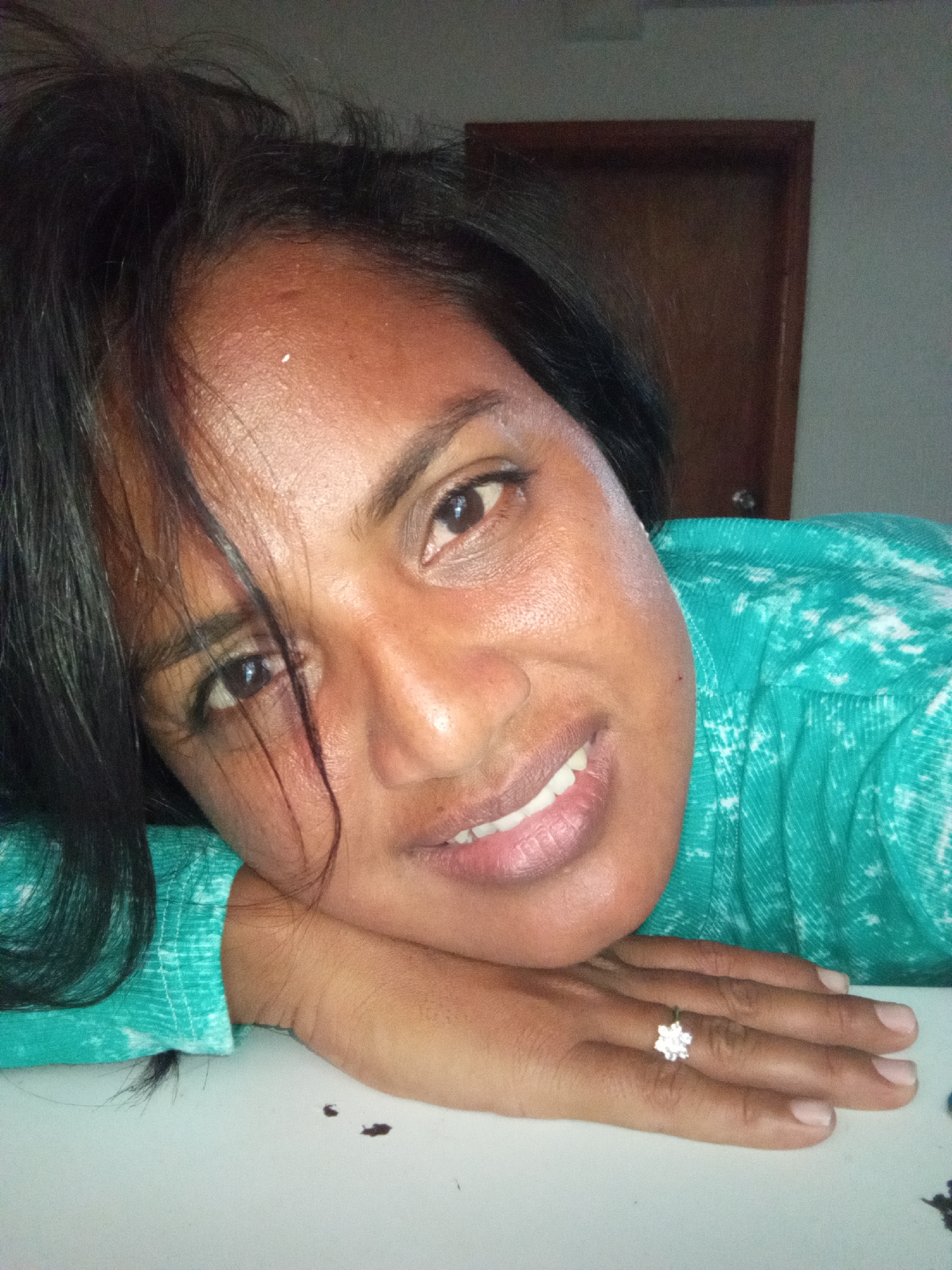A close-up shot shows a woman with dark hair and brown skin, resting her head on her hands. Her eyes are open, and she is smiling, showing her teeth. She is wearing a teal, white, and black patterned shirt and a ring on her right hand. The background is blurred, showing a wooden door and a light-colored wall. The scene appears to be indoors, on a white surface.