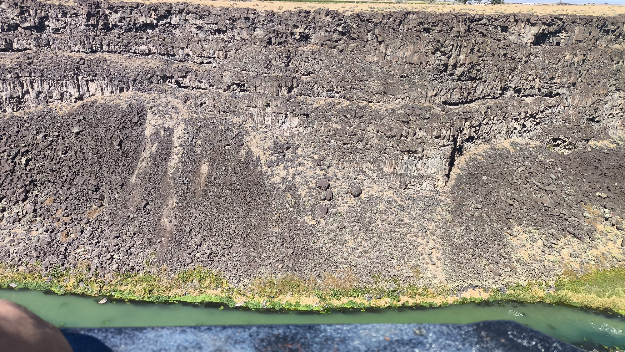 A wide, high-angle drone shot shows a massive, rugged cliff face in Tuttle, United States. The cliff is composed of dark, porous volcanic rock, layered and fractured, with a gentler slope at the bottom transitioning into a sheer drop. Scattered across the rock face are smaller boulders and patches of dry brush. At the base of the cliff, a vibrant green river snakes along, bordered by a narrow strip of lush green vegetation and sandy soil. The sky above is clear and bright. The overall impression is one of raw, natural beauty and geological grandeur.