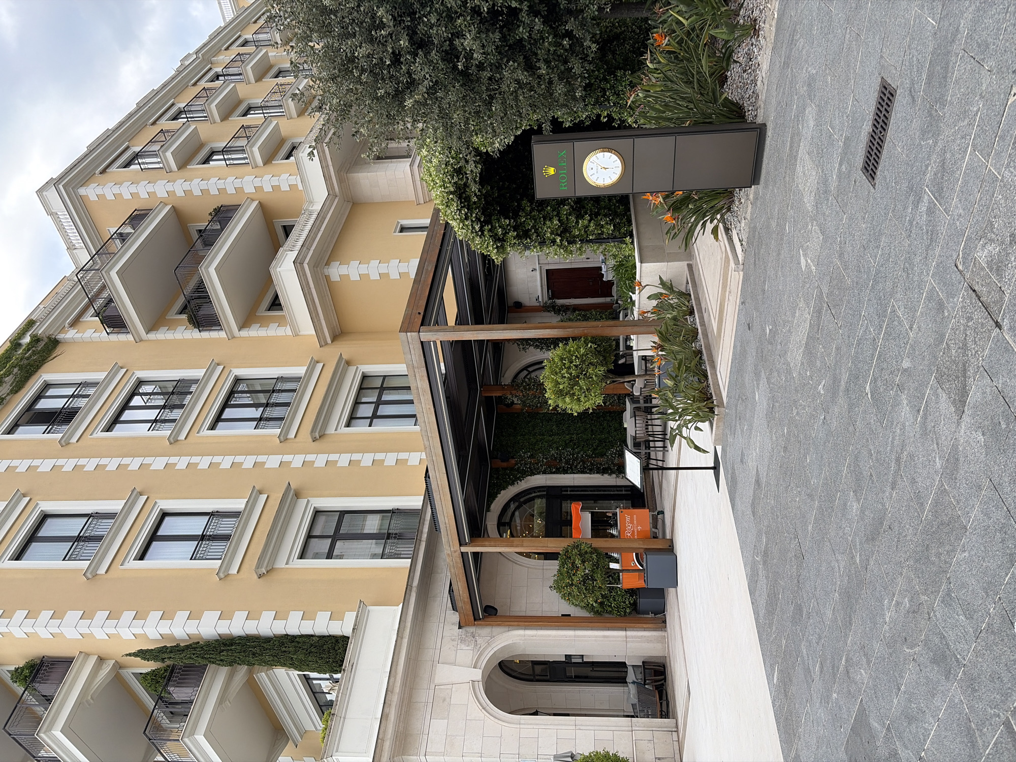 The image depicts the entrance to the Regent Porto Montenegro Hotel in Tivat, Montenegro. The architecture is a beige, multi-story building with black-framed windows and decorative balconies. A wooden pergola structure shades an outdoor seating area in front of the hotel entrance. A Rolex sign with a clock is positioned near the entrance, surrounded by greenery, including trees and flowering plants. The foreground shows a paved stone walkway leading up to the entrance. No people are visible in the scene.