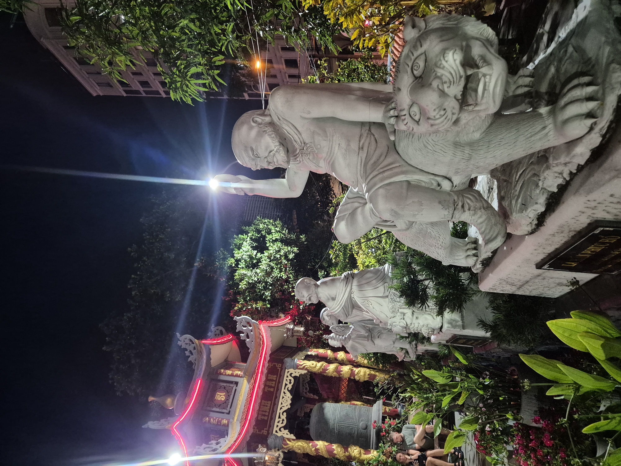 The image shows a night scene in Da Nang, Vietnam, likely near a temple or cultural site. In the foreground, a stone sculpture depicts a bald, bearded man sitting on a tiger. The man is raising his right hand, holding a circular object. Behind him, a line of other stone sculptures are visible, including figures resembling monks or deities. To the left, a traditional Vietnamese pagoda-style building with red neon lighting is illuminated. A large bell hangs in front of the building, supported by ornate golden pillars. Lush green plants and red flowers are arranged around the sculptures and building. In the background, a modern building with multiple windows and trees are visible, along with streetlights. Two individuals, a man and a woman, are seated on a low wall on the left side of the frame, appearing to be observing the scene. The overall atmosphere is dimly lit, with artificial lights from the building and street creating strong highlights and shadows.