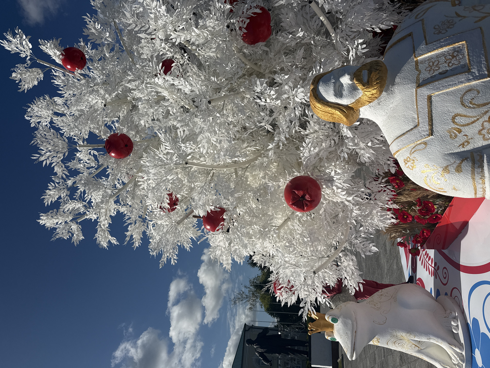 In Dmitrov, Russia, a white artificial tree adorned with red spherical ornaments is showcased outdoors. In the foreground, to the left, a white frog sculpture with green eyes and a golden crown sits on a decorative platform. To the right, a bust of a man with a golden beard and hair, wearing a white robe with golden embellishments, is positioned in front of the tree. Behind the frog and tree, a black silhouette statue of a man stands. The scene is set against a bright blue sky with scattered white clouds, and a building and trees are visible in the background. The ground appears to be paved.