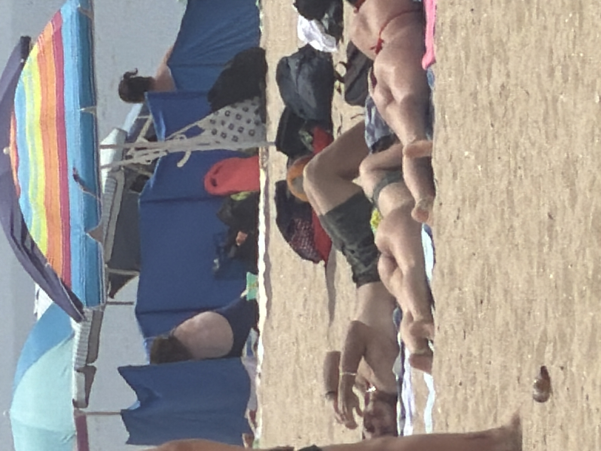 The image shows a beach scene in Espinho, Portugal. Several people are lying on the sand, some under beach umbrellas. In the foreground, a person is lying on their back with their arms raised. To the right, another person is lying on their stomach with their legs and buttocks visible. In the background, two beach umbrellas, one blue and one with rainbow stripes, provide shade. Beach tents and bags are scattered on the sand. The ocean is visible in the distance.