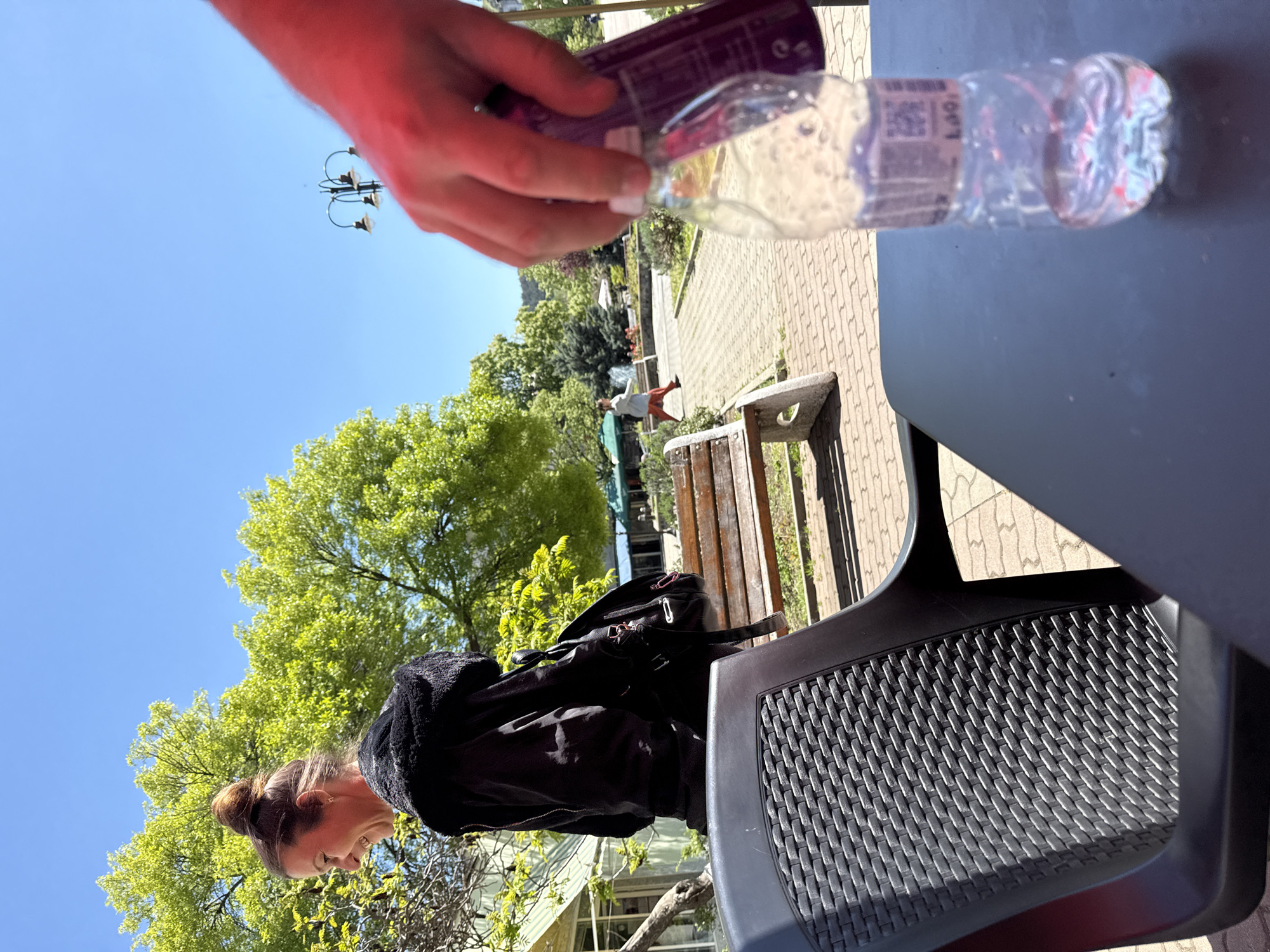 The image shows an outdoor scene with a woman sitting in a black plastic chair at a table on a patio or outdoor area. The woman is wearing a black jacket and has her hair up. A black backpack is on the bench behind her. A person's hand is holding a water bottle over the table. The background includes trees, blue sky, a brick walkway, a bench, and another person walking in the distance. The location appears to be a park in Karnobat, Bulgaria.