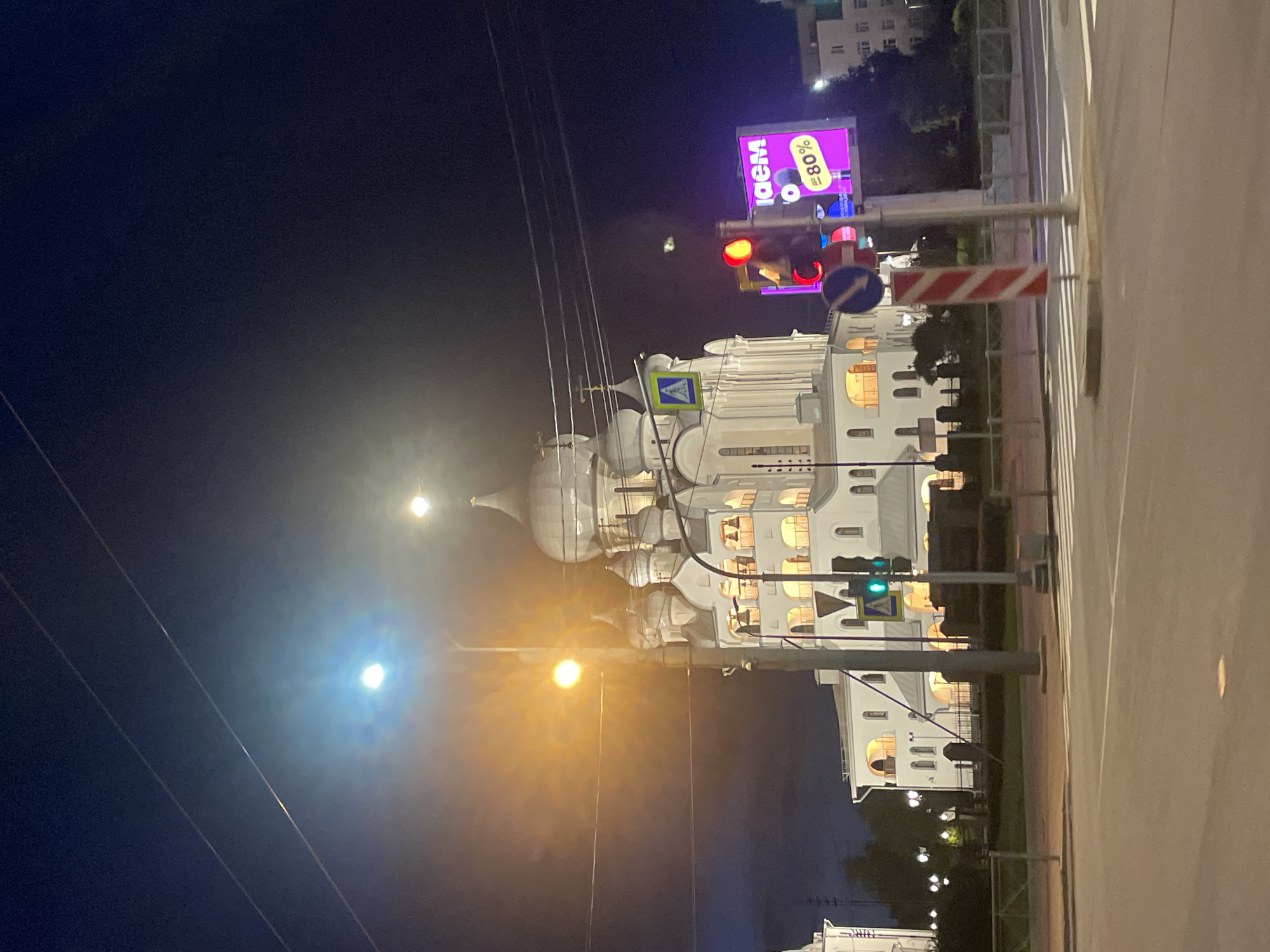 The image displays a vibrant night scene of a city street, featuring a large, ornate white building and various urban elements. The scene appears to be rotated 90 degrees counter-clockwise.
The central subject is a majestic, multi-domed white building, likely a church or cathedral, distinguished by its intricate architecture and numerous arched windows glowing with warm interior light. Multiple domes with subtle gold-colored crosses are visible at the top.
In the foreground and midground, a dark asphalt road with clear white pedestrian crossing stripes and lane markings is present. The night sky is dark blue, illuminated by a mix of artificial lights. Bright streetlights cast distinct glows; one has a warm yellow hue, and another emits a cooler white-blue light, both creating prominent lens flares.
Several street furniture elements are visible, including traffic lights, one displaying a red signal and another a green signal. Various road signs are also present: a blue square sign with a white pedestrian icon, a blue circular sign with a white arrow pointing left, and a red and white striped barrier or pole. Overhead, multiple thin power lines crisscross the frame.
High on the right side of the image, a large, brightly illuminated purple LED billboard is prominent. The visible text on this billboard reads "IDEM" (likely Cyrillic for "ИДЁМ") and "до -80%", indicating a commercial advertisement for a discount. No people are visible in the scene.