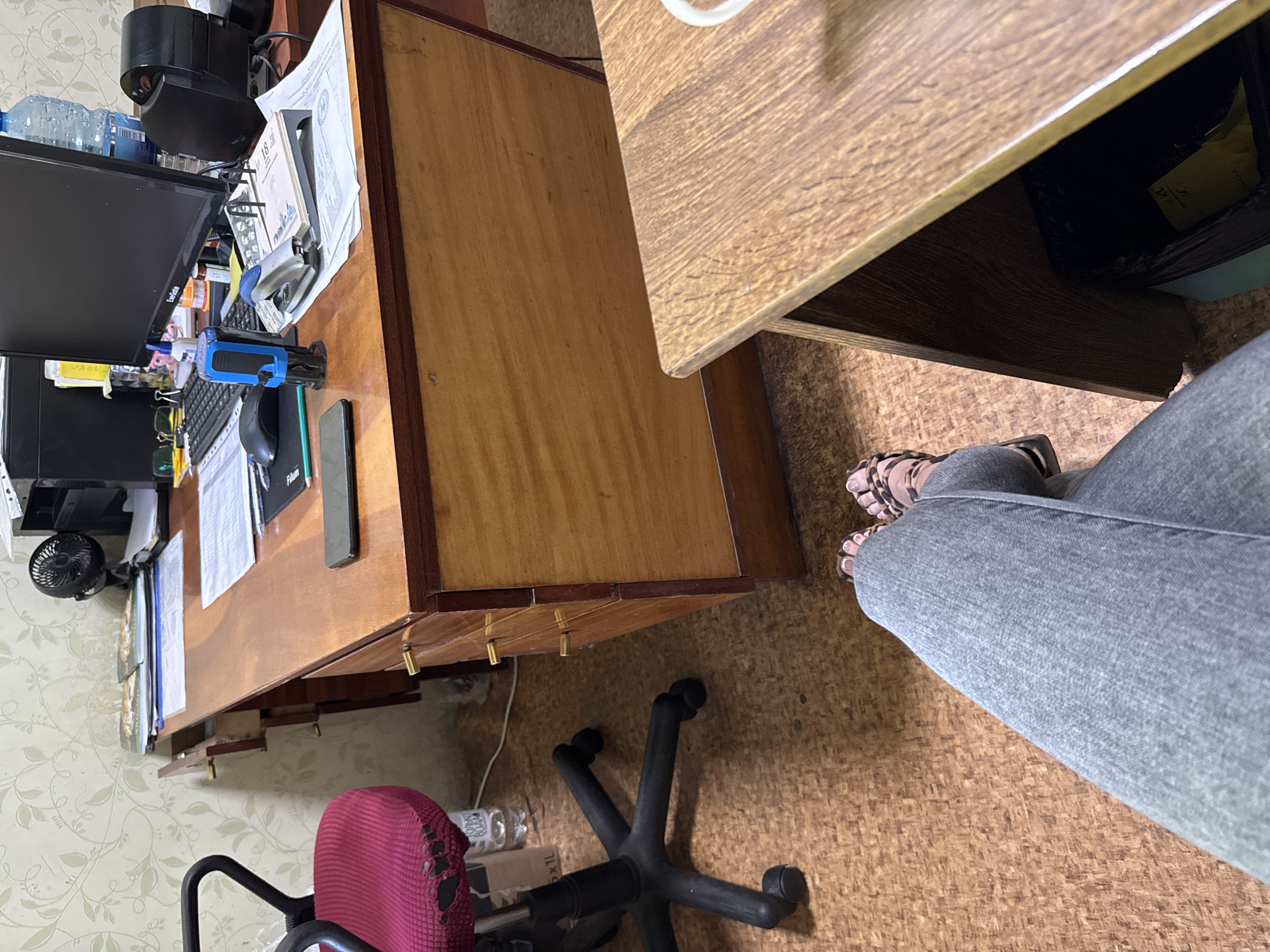 The media file shows an office desk from a first-person perspective. The desk is made of wood and has several objects on it, including a computer monitor, keyboard, a blue and black stamp, papers, a phone, and a small fan. There is also a trash can under a second desk, which is made of a lighter wood. The floor is covered with a cork-like material. The background shows a patterned wallpaper. The image is taken from a seated position, and the legs of a person wearing grey jeans and leopard-print sandals are visible in the foreground. The lighting suggests it is daytime, but there are no clear indicators of the weather or a specific location. The scene appears to be a home office or a personal workspace. There is some text visible on the papers, but it is not legible enough to be read.