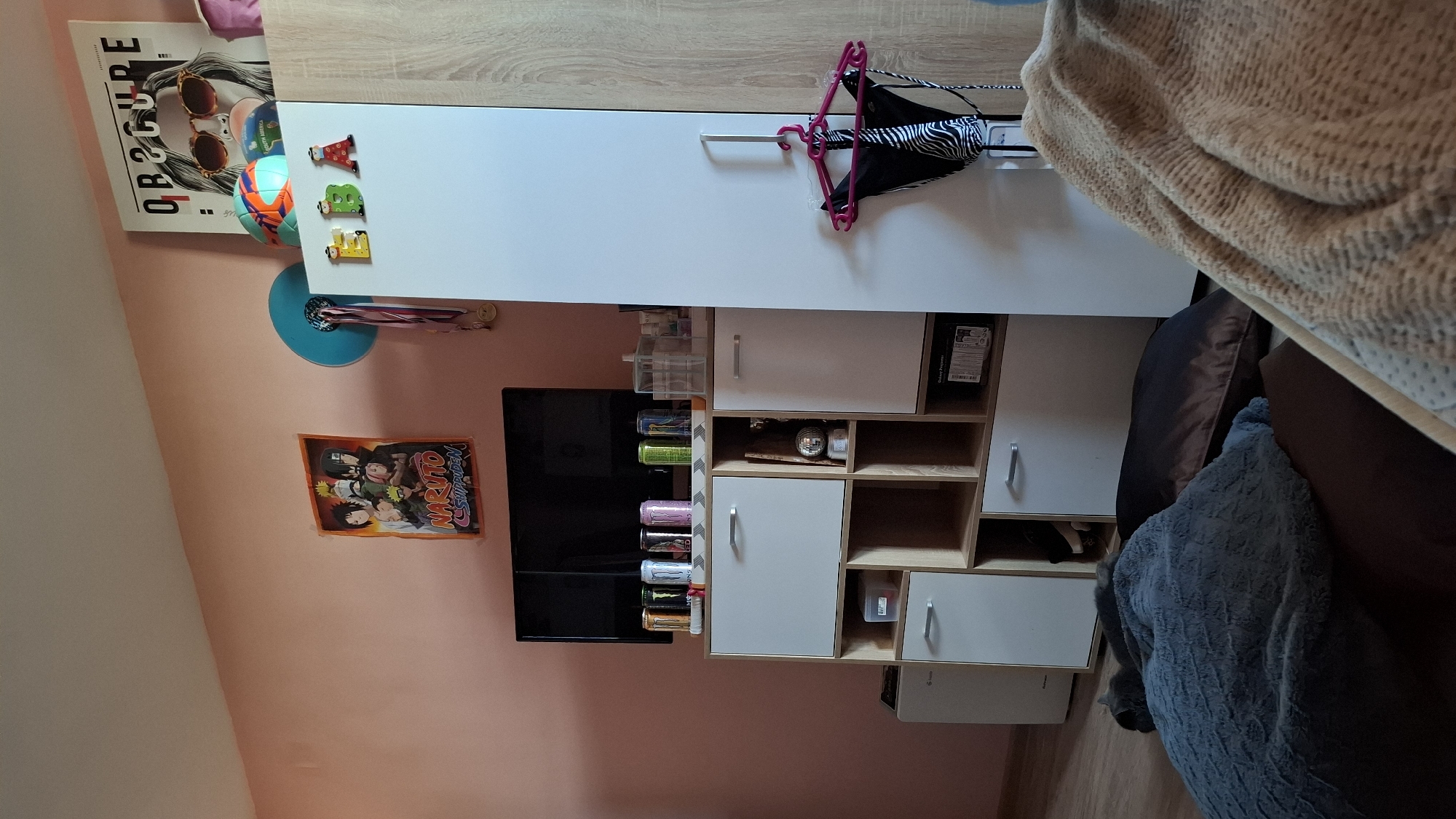 The image shows a cluttered interior space, possibly a teenager's room. A white cabinet with drawers and open shelves occupies the center of the frame, partially obscuring a television mounted on the wall. Various items are placed on and within the cabinet, including containers, cans of energy drinks, a hard drive, and decorative objects. A zebra-print bikini top hangs on a pink hanger attached to the cabinet's side. On the wall, a poster featuring an image of a woman wearing sunglasses is visible above the cabinet, along with a soccer ball attached to the cabinet's side with magnets, and a blue disc with hanging items below. A "Naruto Shippuden" poster is affixed to the wall below the TV. A brown sofa with pillows and a blue blanket sits to the right of the cabinet. The walls are painted in a light peach color. Based on other contextual cues, the location is likely Sofia, Bulgaria. No people are visible in the image. The scene depicts a typical interior shot of a furnished room with personal belongings.