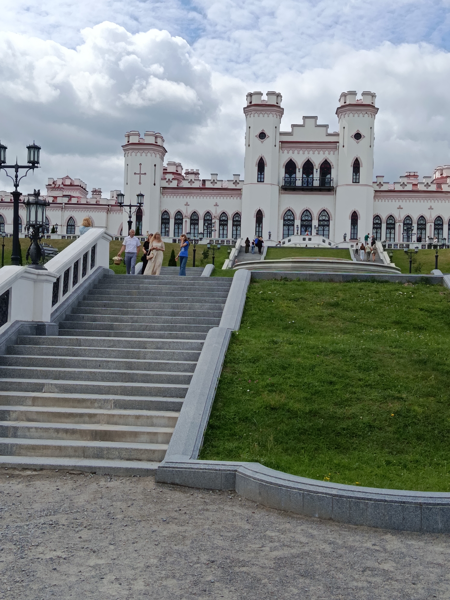 The image shows a bright white castle-like building with red trim and turrets under a cloudy sky. In the foreground, a wide stone staircase leads up towards the building. Several people are walking up the stairs, including a man carrying a basket of flowers and a woman in a long dress. To the right of the stairs is a green, grassy slope. The location is identified as the Pushkin Museum in Kvasovichi village, Kvasevitski Selski Sovet, Belarus. The scene depicts a sunny but overcast day, with people visiting or exiting the historical site.