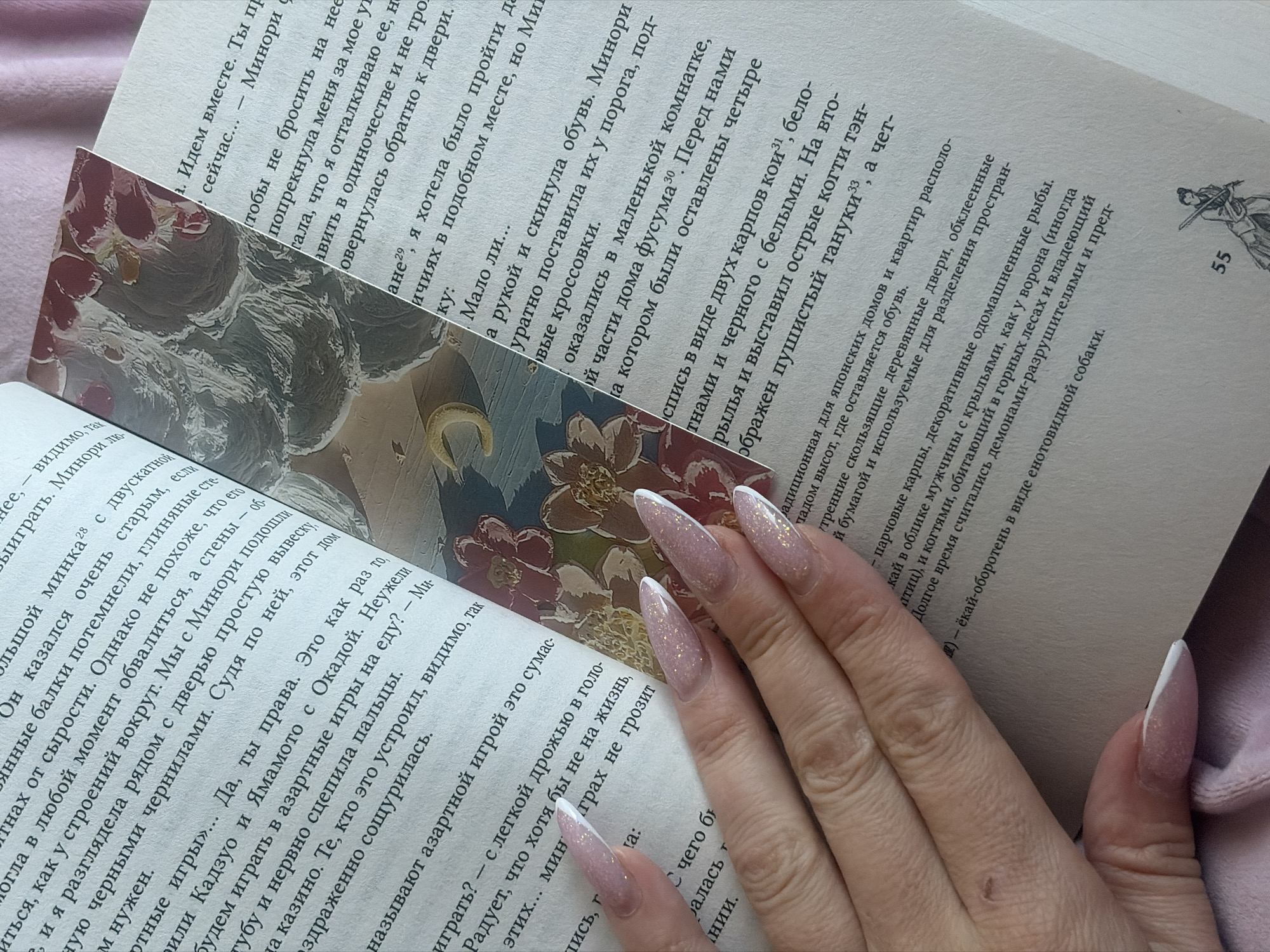The image displays a close-up of a person reading a book, resting on a soft pink fabric background, likely in Saint Petersburg, Russia. A hand with long, almond-shaped nails, featuring glittery pink and white French tips, holds the book open. A decorative bookmark, depicting a stylized figure with a golden halo and abstract patterns, possibly representing water or clouds, with floral accents, is placed between the pages.
The book is written in Russian, and visible text includes character names and terms related to Japanese culture and folklore. Keywords such as "Минори" (Minori), "фусума" (fusuma - Japanese sliding doors), "карпов кои" (koi carp), "тэн-гу" (tengu - a mythical creature), "тануки" (tanuki - another mythical creature), and "ёкай-оборотень" (yokai-shapeshifter) are discernable, suggesting the book's narrative explores Japanese mythology or history. Footnote numbers (e.g., 28, 29, 30, 31, 33) and the page number "55" are visible.
The scene captures a moment of quiet indoor activity, emphasizing leisure and personal style through the distinct nail art and the unique bookmark. No specific time of day or weather conditions are discernible.